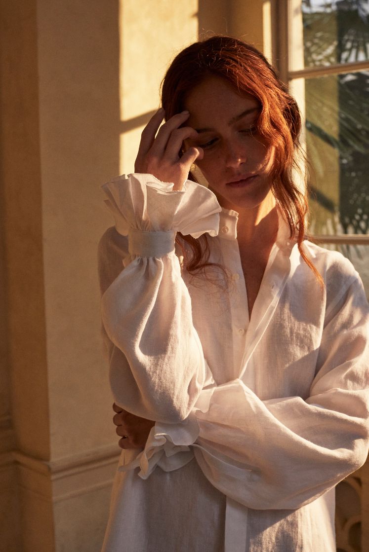 Photo plan américain d'une femme rousse portant un chignon, deux mèches tombantes sur son visage. Elle porte une chemise BOUDOIR dans un lin blanc plein de détails raffinés : un col haut subtilement bordé de petits volants pour une touche de sophistication et des manches légèrement bouffantes ornées de volants, qui tombent avec élégance sur la main. Sa main touche délicatement son visage, l'air pensive. La chemise confectionnée dans un lin belge de Libeco.