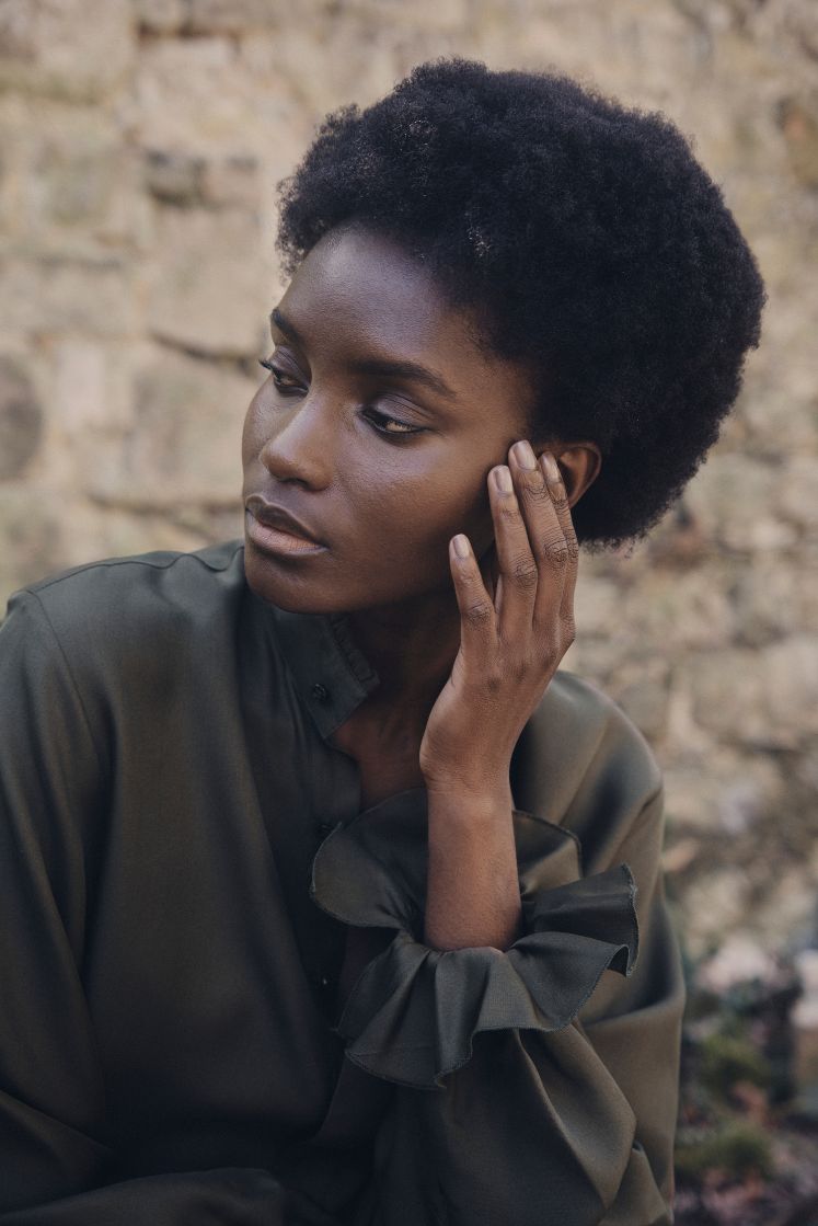 Femme assise portant la chemise BOUDOIR en soie couleur kaki avec poignets à volants, col ancien et coupe fluide, posant devant un mur de pierre.
