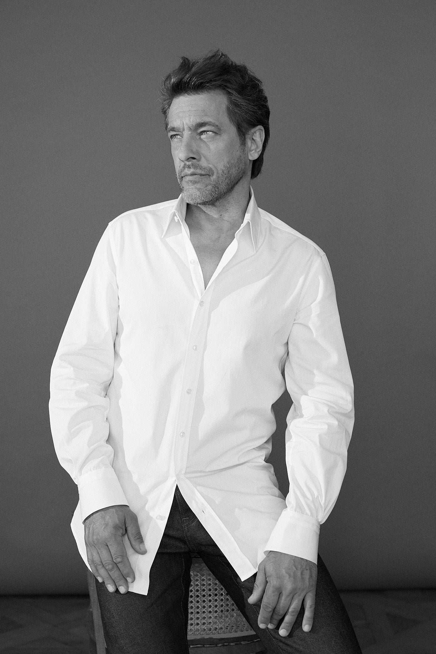 Photo noir et blanc plan américian d'un homme portant une chemise en popeline blanche double retors, col français et poignets bourrienne et un jean. L'homme est debout, légèrement assis à une chaise derrière lui. Ses mains sont posées sur ses jambes, il tourne son regard à sa droite.