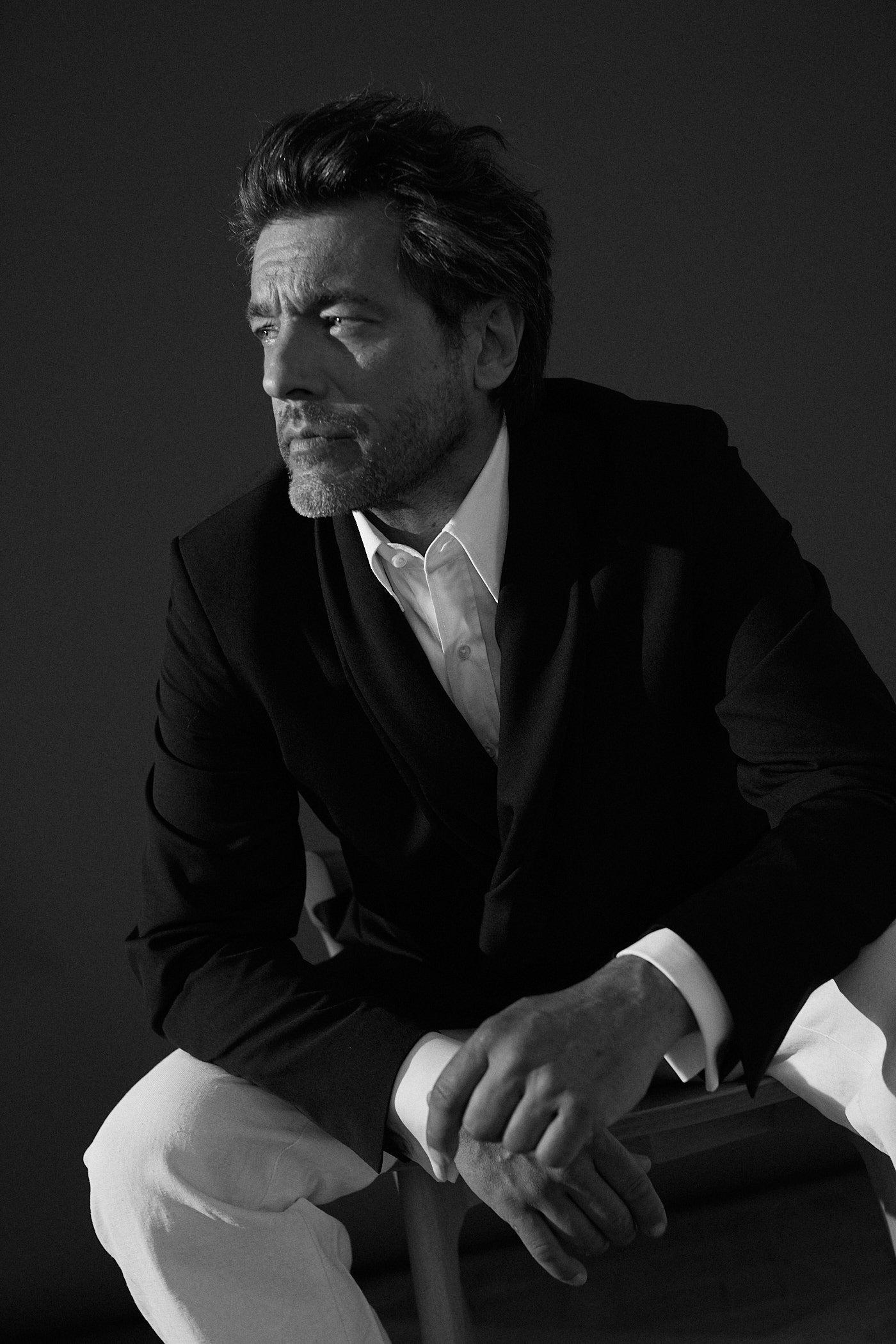 Photo noir et blanc plan américain d'un homme assis qui regarde à sa droite, ses deux mains se touchent. Il porte une chemise blanche en popeline blanche double retors, poignets mousquetaires et col français ert une vers de costard noire avec un pantalon de costume blanc.