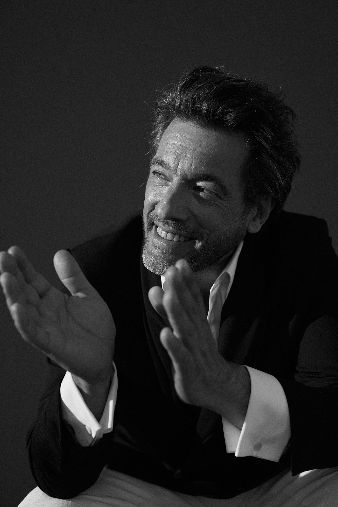 Photo noir et blanc plan serré d'un homme assis ses deux coudes sont posées sur ses genous, ses deux mains sont levées et vont vers sa droite. Il porte une chemise en popeline blanche double retors, poignets mousquetaires et col français avec une veste costard noire. Il regarde à sa droite, il sourit.