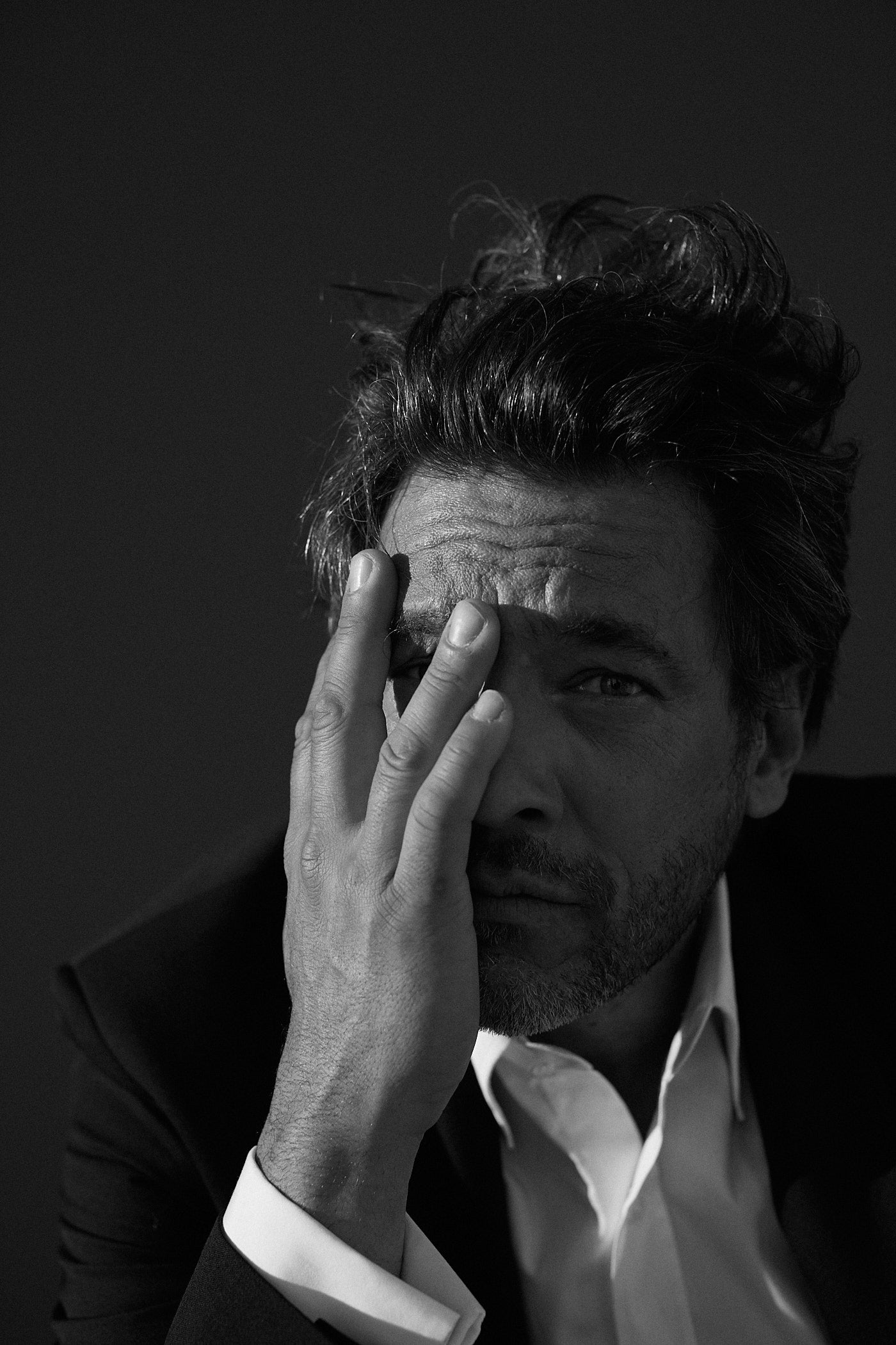 Photo noir et blanc plan serré d'un homme qui regarde l'objectif. Il tient sa main droite sur son visage et cache un oeil. Il porte une chemise blanche en popeline blanche double retors, poignets mousquetaires et col français.