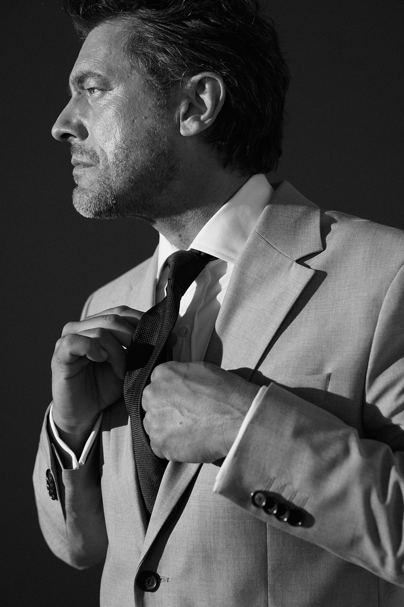 Photo plan américain en noir et blanc d'un homme en costard, son visage est de profil il regarde à gauche. Il porte un blazer, une chemise avec un col italien et une cravate noire. Ses mains touchent les boutons du haut de sa chemise, par dessous sa cravate.