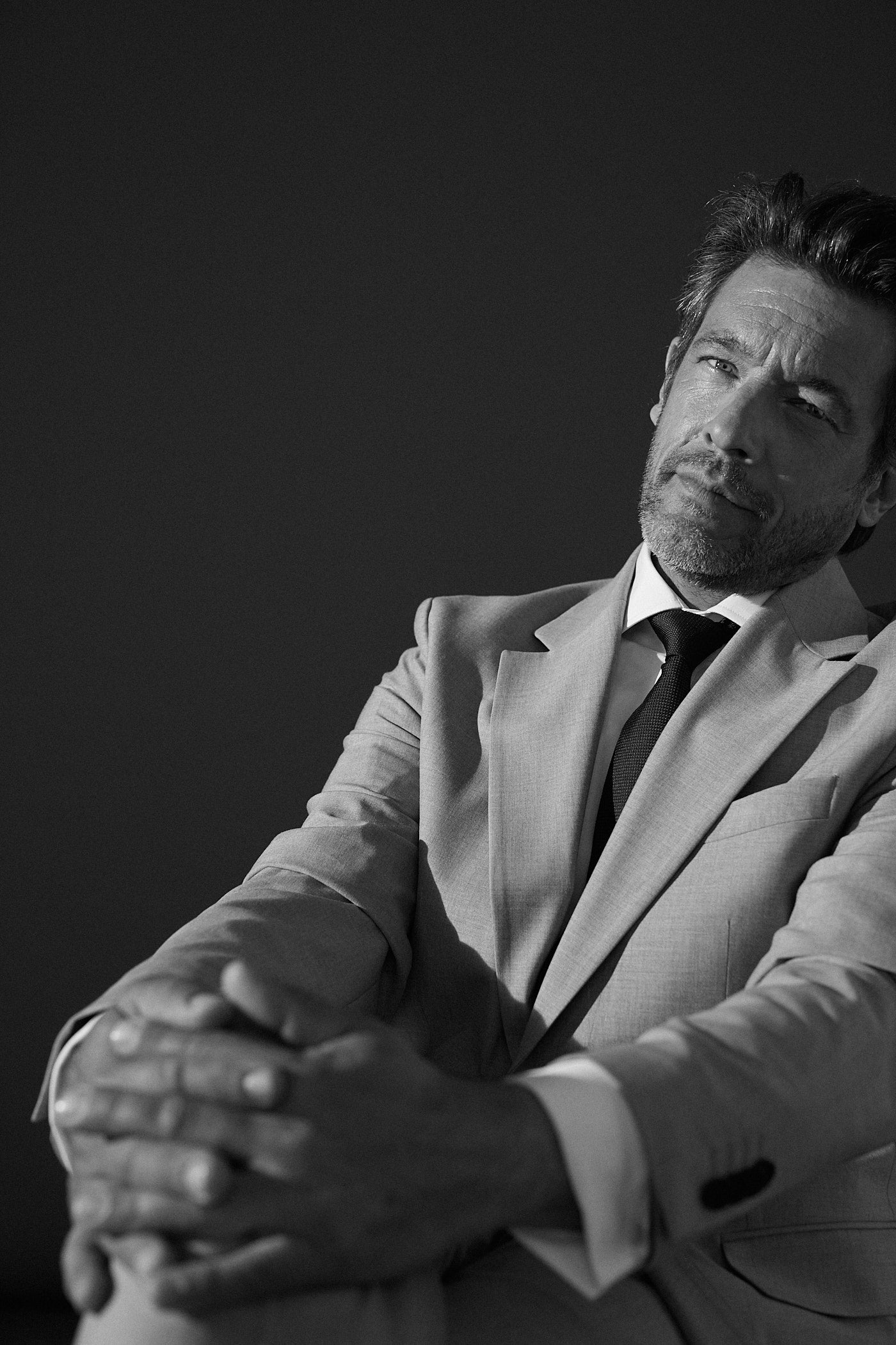 Photo en noir et blanc d'un homme assis en costard beige. Il croise ses jambes et tient avec ses mains également croisé, son genou droit. Il porte en dessous de son blazer une chemise blanche col italien et une cravate noire. Le visage de l'homme regarde l'objectif avec un regard profond.