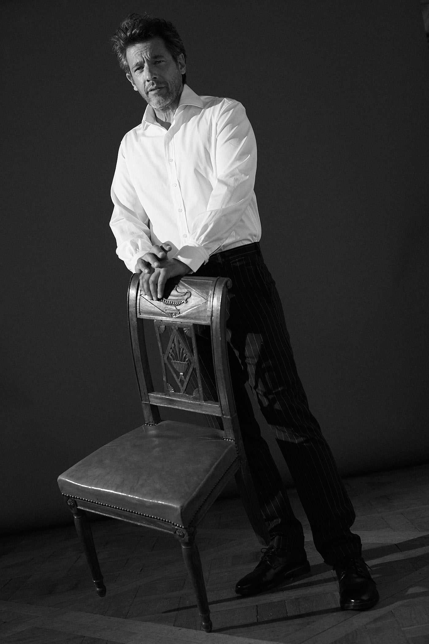 Photo plan américain en noir et blanc. Un homme est debout il touche avec ses mains une chaise devant lui qui bouscule vers l'avant. Il porte une chemise blanche en popeline double retors, col italien avec des poignets Bourrienne, un pantalon de tailleur noir avec des rayures blanches. L'homme regarde devant l'objectif.