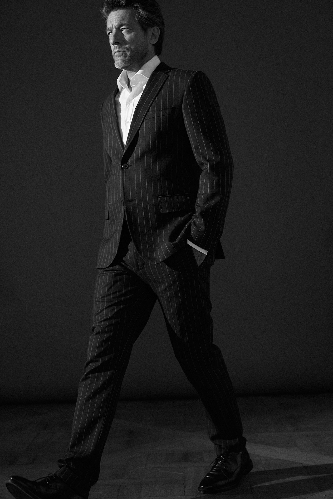 Photo en noir et blanc, plan moyen, un homme sur un fond gris portant un costard noir avec des rayures blanches. Il porte en dessous une chemise blanche col italien en popeline blanche double retors. Sa main gauche est à l’intérieur de son tailleur, il est entrain de marcher légèrement incliné vers la gauche.