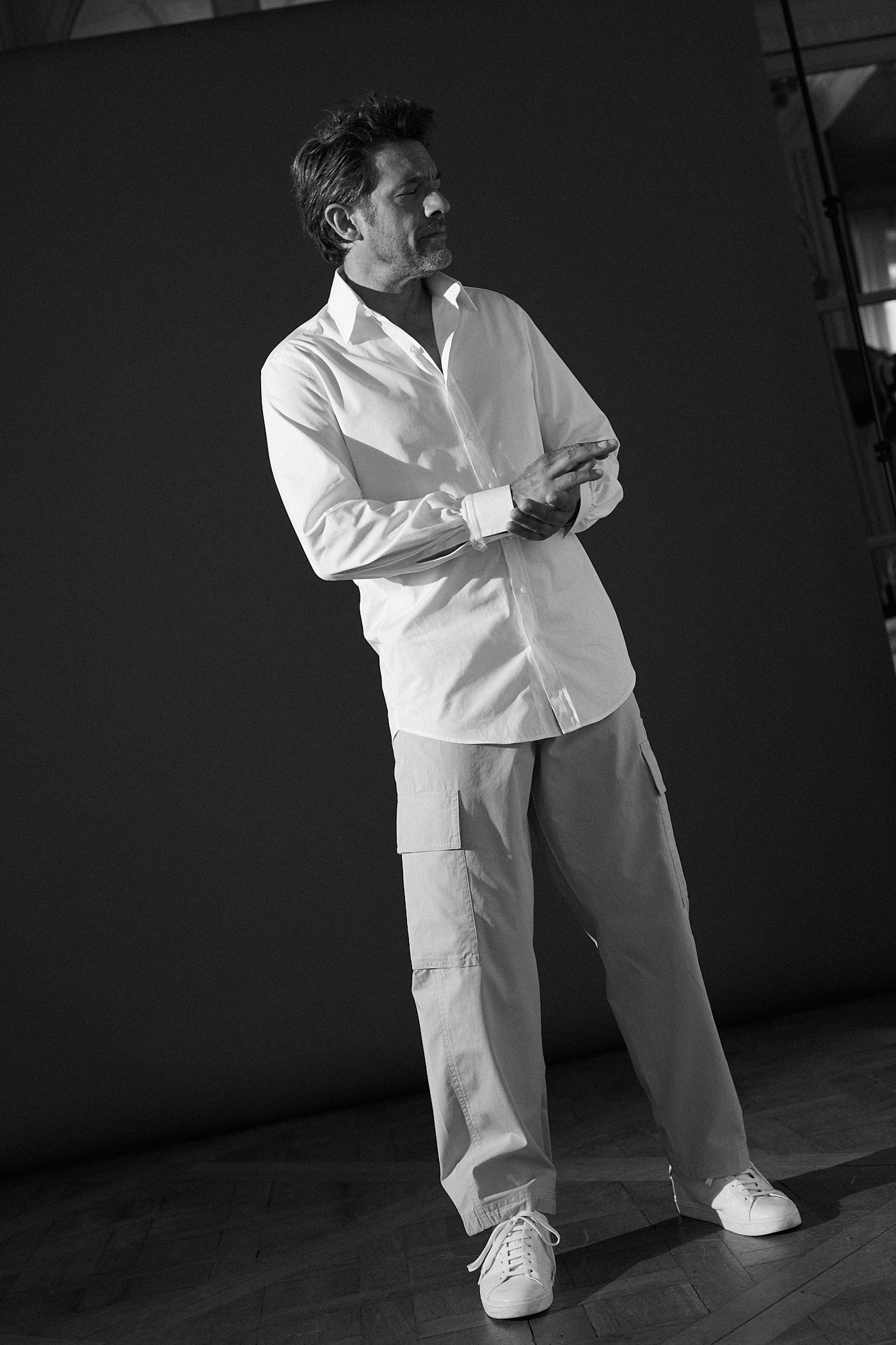 Photo noir et blanc plein pied d'un homme portant une chemise en popeline blanche double retors, col français et poignets bourrienne et un pantalon beige avec deux poches à l'avant. L'homme est debout il tient avec sa main gauche son autre main qui touche le poignet. Son visage est de profil.