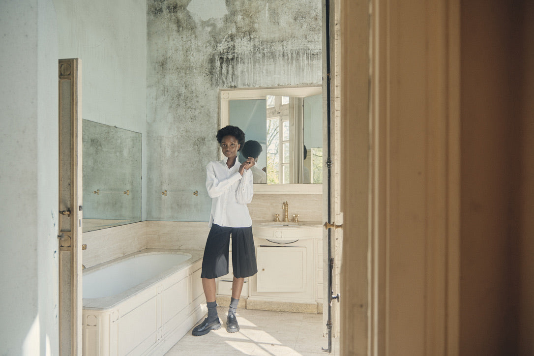 Femme dans une salle de bain lumineuse, vêtue d'une chemise blanche Bourrienne Paris X, d'un bermuda noir, de mocassins noirs avec chaussettes grises.