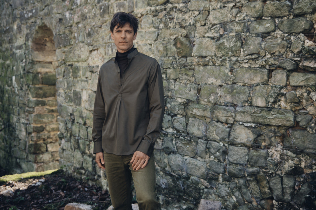 Homme debout devant un mur de pierre ancien, vêtu de la chemise ÉPICURIEN Kaki en popeline de coton (coupe Bourrienne ample, col officier biseauté, poignets classiques), portée sur un col roulé noir et un pantalon kaki, pour une allure décontractée et structurée.