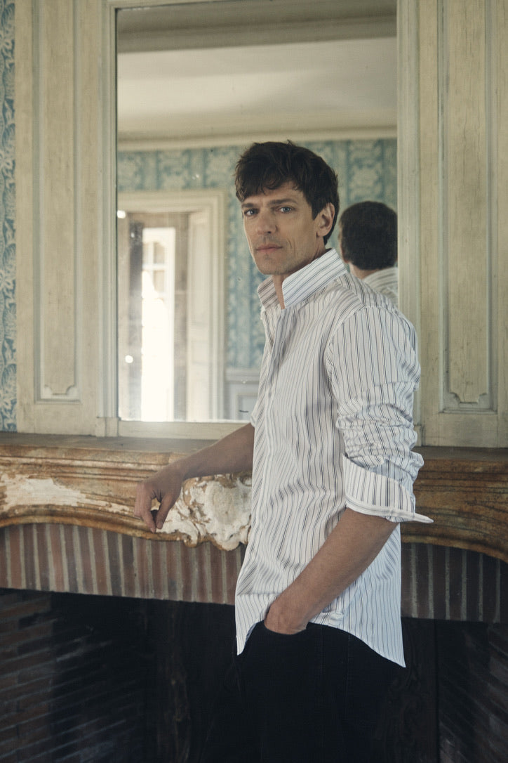 Un homme de profil trois-quarts, main droite posée sur le rebord de la cheminée et main gauche glissée dans la poche de son pantalon sombre, arbore une chemise ajustée en popeline kaki clair à fines rayures verticales blanches et gris clair. Le col montant, ouvert au deuxième bouton, et les manches légèrement retroussées dévoilent un poignet fin orné d’un bracelet de manche boutonné par un petit bouton nacré. Le tissu fluide crée un subtil jeu de plis, mis en valeur par une lumière douce qui accentue la te