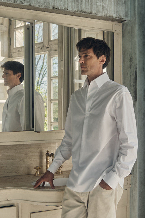 Une photo plan poitrine d'un homme qui est debout, sa main gauche rentré dans son pantalon et l'autre posé sur le rebord d'un évier de salle de bain. Il regarde de profil, il porte une chemise 100% Coton, fabriqué en Europe, une coupe Bourrienne, un col Français et des Poignets Bourrienne.