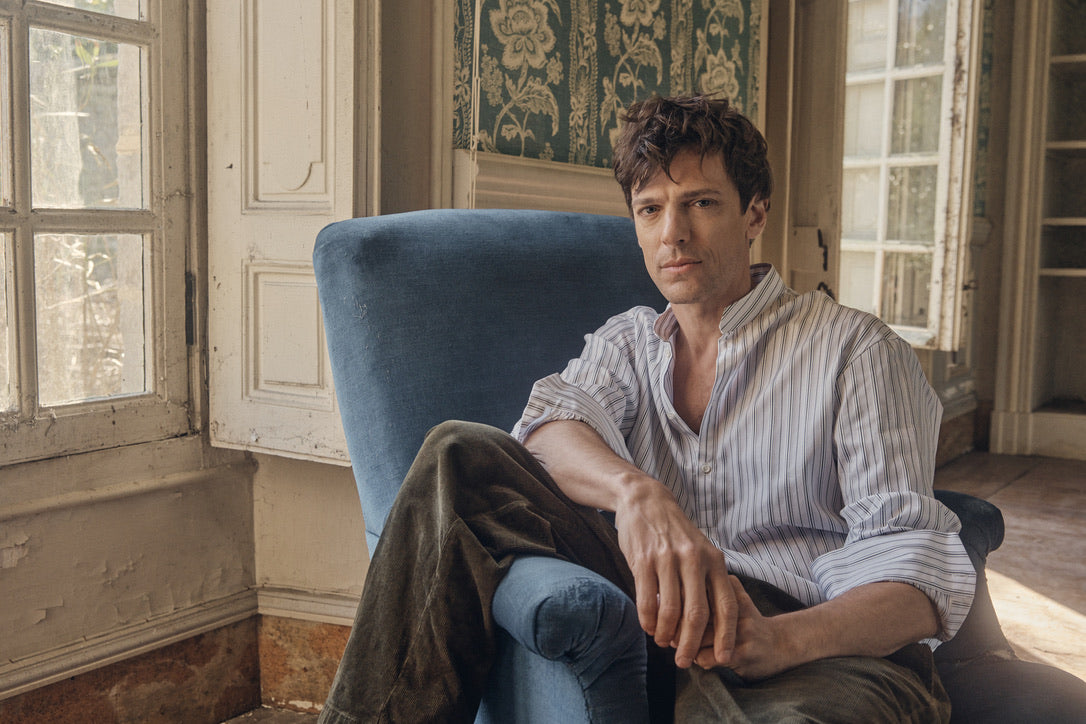 Homme assis dans un fauteuil bleu face à l’objectif, vêtu de la chemise ÉPICURIEN Rayée Kaki en popeline de coton (coupe Bourrienne ample, col officier biseauté, poignets classiques retroussés), dans un intérieur ancien baigné de lumière.