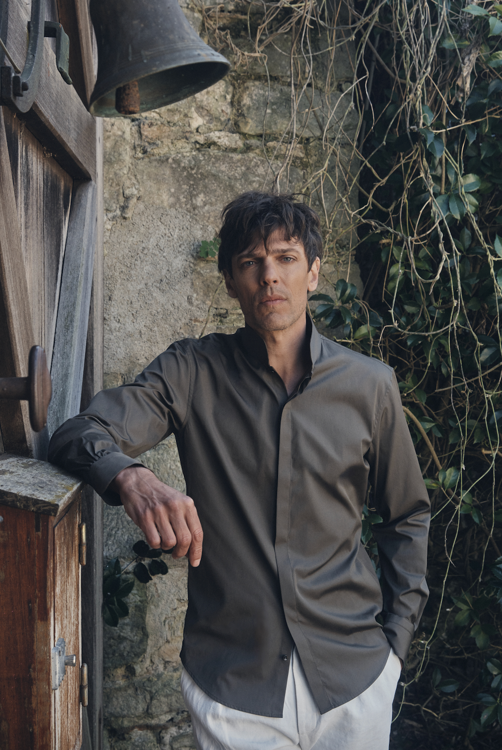 Un homme de face, adossé à un mur de pierre et une porte en bois patiné sous une cloche en métal, le regard pensif vers l’objectif. Il porte une chemise ajustée kaki foncé en popeline au col montant ouvert et manches longues boutonnées aux poignets, associée à un pantalon blanc cassé taille haute. Le contraste entre la texture brute du décor et la coupe épurée de la tenue dégage une allure à la fois naturelle et sophistiquée.