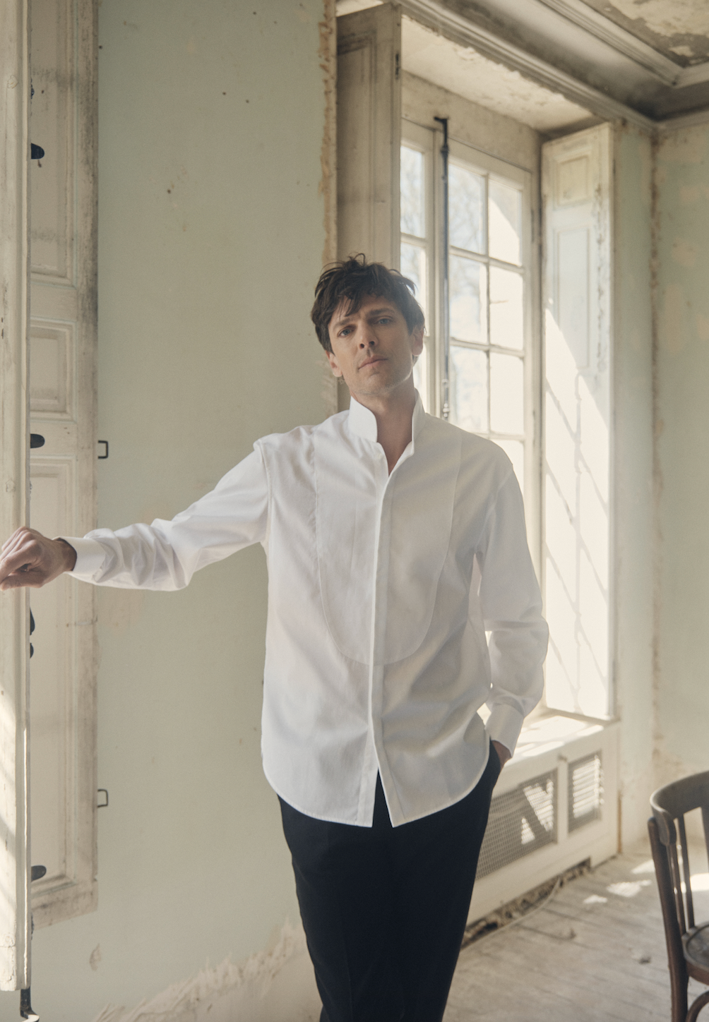 Photo plan américain d'un homme qui se tient debout, le visage face à la caméra, sa main gauche rentrée dans son pantalon, l'autre posé sur une fenêtre. Il porte une chemise blanche, coupe Bourrienne, poignets Français, col haut officier avec un plastron.