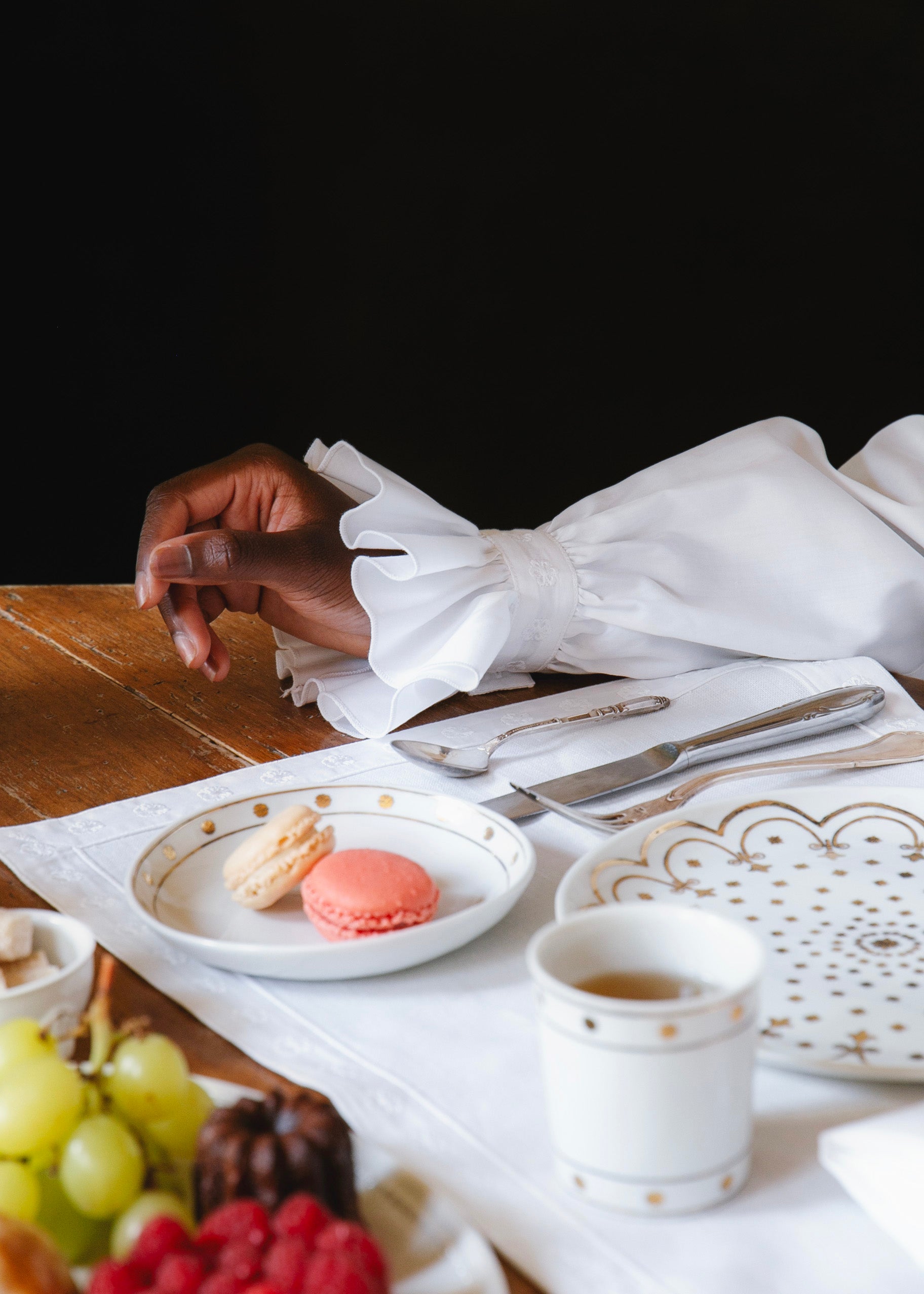 On aperçoit le poignet à volants de la chemise EXQUISE, posé sur une table dressée avec des macarons, des fruits et des pâtisseries.