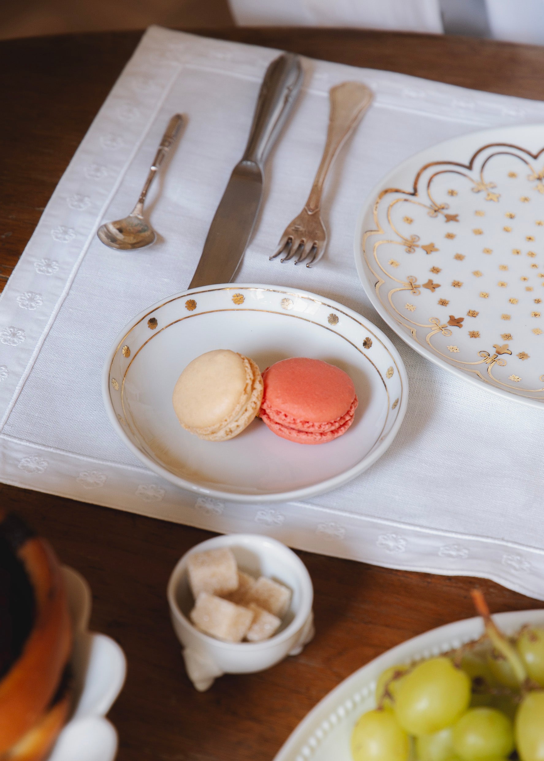 Table dressée avec la coupelle en porcelaine blanche aux motifs dorés qui contient deux macarons, l'assiette également ornée de motifs dorés reprenant la fleur de l'Hôtel de Bourrienne, et le set de table brodé du même motif.