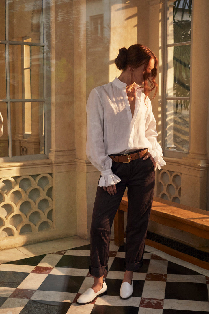 Photo en plan plein pied d'une femme rousse avec un chignon et deux mèches qui tombent sur son visage. Son visage est tourné à sa gauche, elle porte une chemise BOUDOIR en LIN Blanc aérien et léger. Des détails raffinés sur le col haut subtilement bordé de petits volants et des manches légèrement bouffantes ornées de volants, qui tombent avec élégance sur la main. Elle porte un pantalon léger marron, une ceinture en cuir marron et des mocassins en cuir blancs.