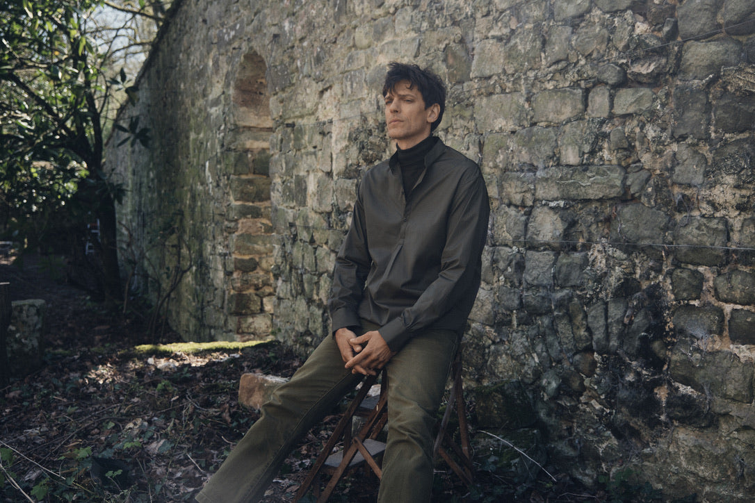 Homme assis sur un tabouret devant un vieux mur en pierre, vêtu de la chemise ÉPICURIEN Kaki en popeline de coton (coupe Bourrienne ample, col officier biseauté, poignets classiques), superposée à un col roulé noir et un pantalon kaki, dans un jardin sec aux feuilles tombées.