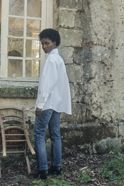 Femme portant la chemise MERVEILLEUSE, posant devant la fenêtre d'une ancienne maison.
