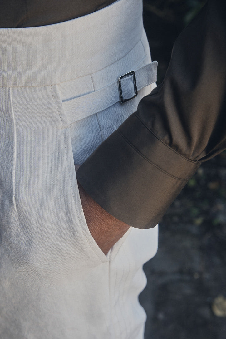 Gros plan sur la taille haute du pantalon blanc cassé, où l’on distingue une mèche fine de tissu pris dans une boucle métallique argentée pour ajuster la largeur. La poche latérale accueille délicatement le poignet droit de la chemise kaki foncé, contrastant nettement la finesse du tissage clair et la texture satinée du tissu sombre.