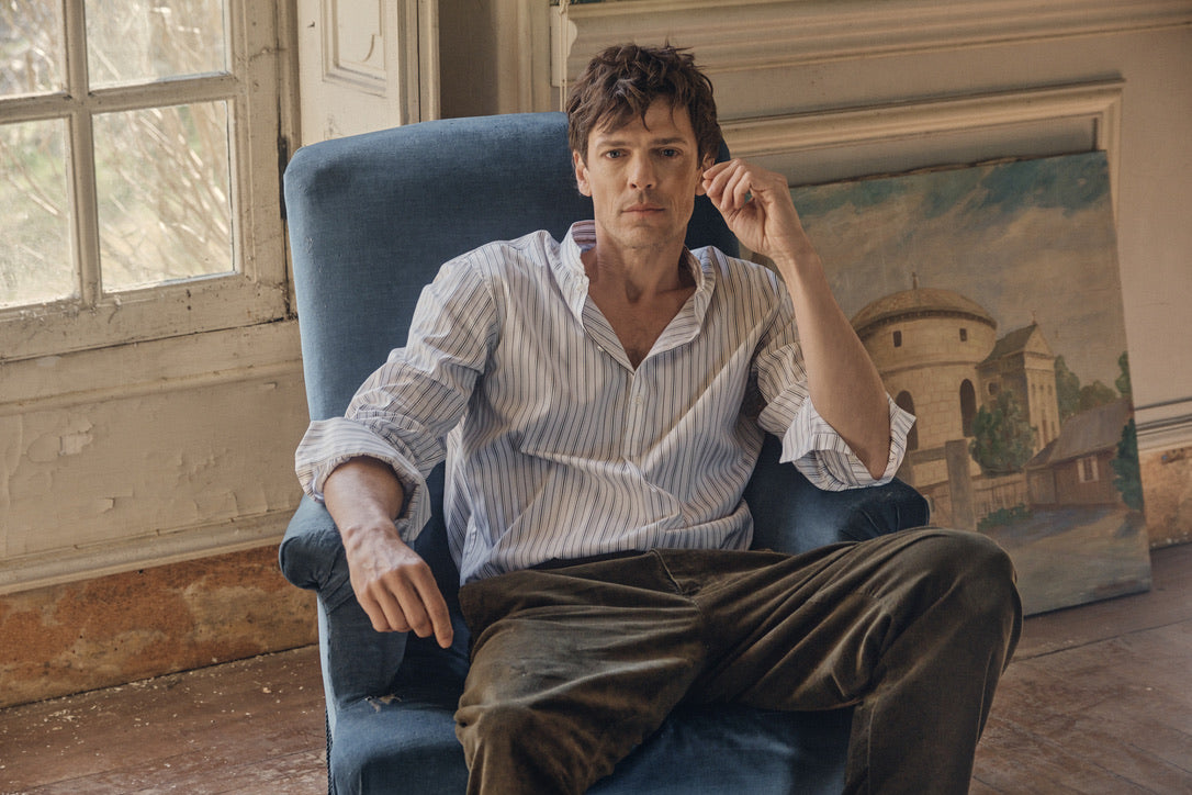 Homme assis dans un fauteuil bleu en velours, portant la chemise ÉPICURIEN Rayée Kaki en popeline de coton (coupe Bourrienne ample, col officier biseauté, poignets retroussés) et un pantalon kaki, dans un intérieur ancien baigné de lumière naturelle