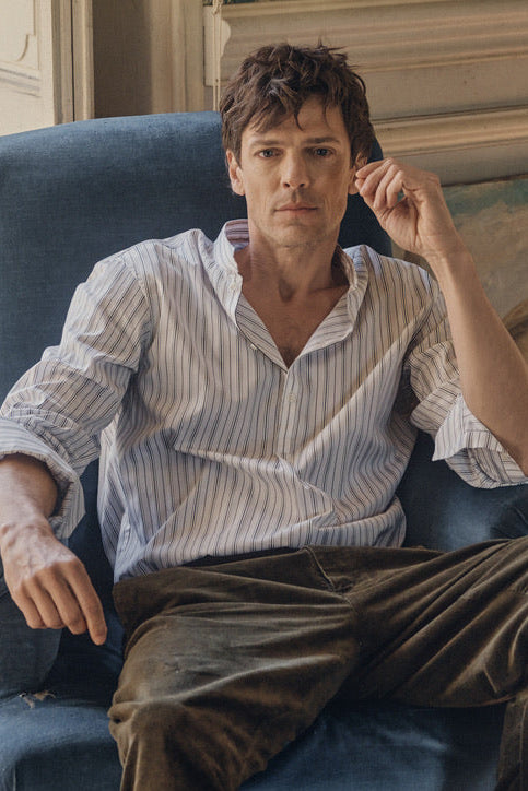 Homme assis dans un fauteuil bleu en velours, portant la chemise ÉPICURIEN Rayée Kaki en popeline de coton (coupe Bourrienne ample, col officier biseauté, poignets retroussés) et un pantalon kaki, dans un intérieur ancien baigné de lumière naturelle