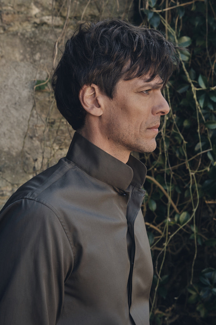 Profil latéral d’un homme vêtu d’une chemise en popeline kaki foncé à col montant, le premier cran ouvert, manches longues ajustées sur les avant-bras. En arrière-plan, un mur de pierre grise et un rideau de lierre créent un contraste naturel avec la coupe épurée de la chemise.