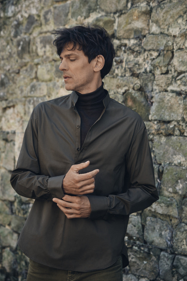 Homme debout devant un mur de pierre, ajustant les poignets de sa chemise ÉPICURIEN Kaki en popeline de coton : coupe Bourrienne ample, col officier biseauté, poignets classiques, portée sur un col roulé noir pour une allure contemporaine et structurée.