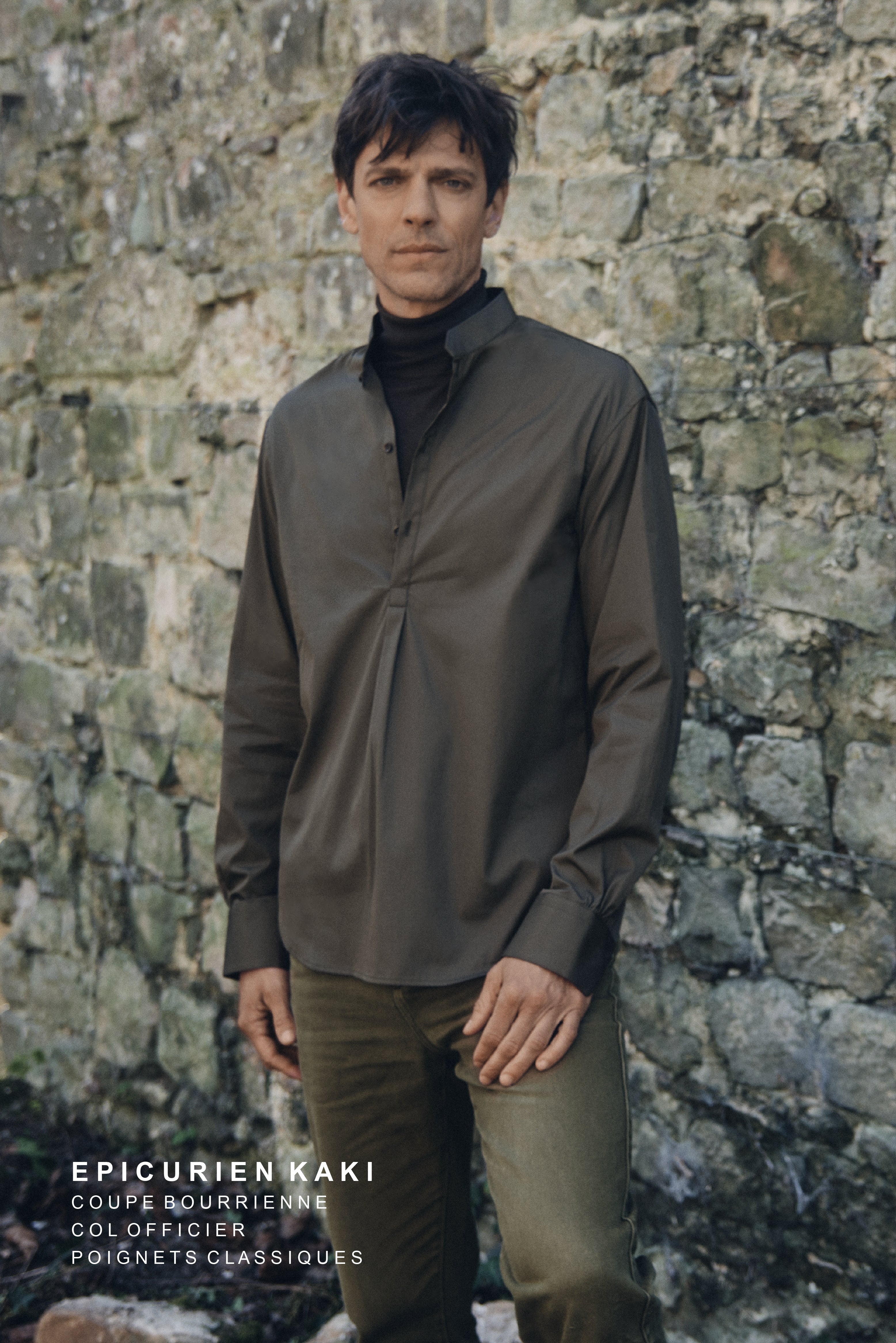 Homme debout devant un mur de pierre ancien, vêtu de la chemise ÉPICURIEN Kaki en popeline de coton (coupe Bourrienne ample, col officier biseauté, poignets classiques), portée sur un col roulé noir et un pantalon kaki, pour une allure décontractée et structurée.