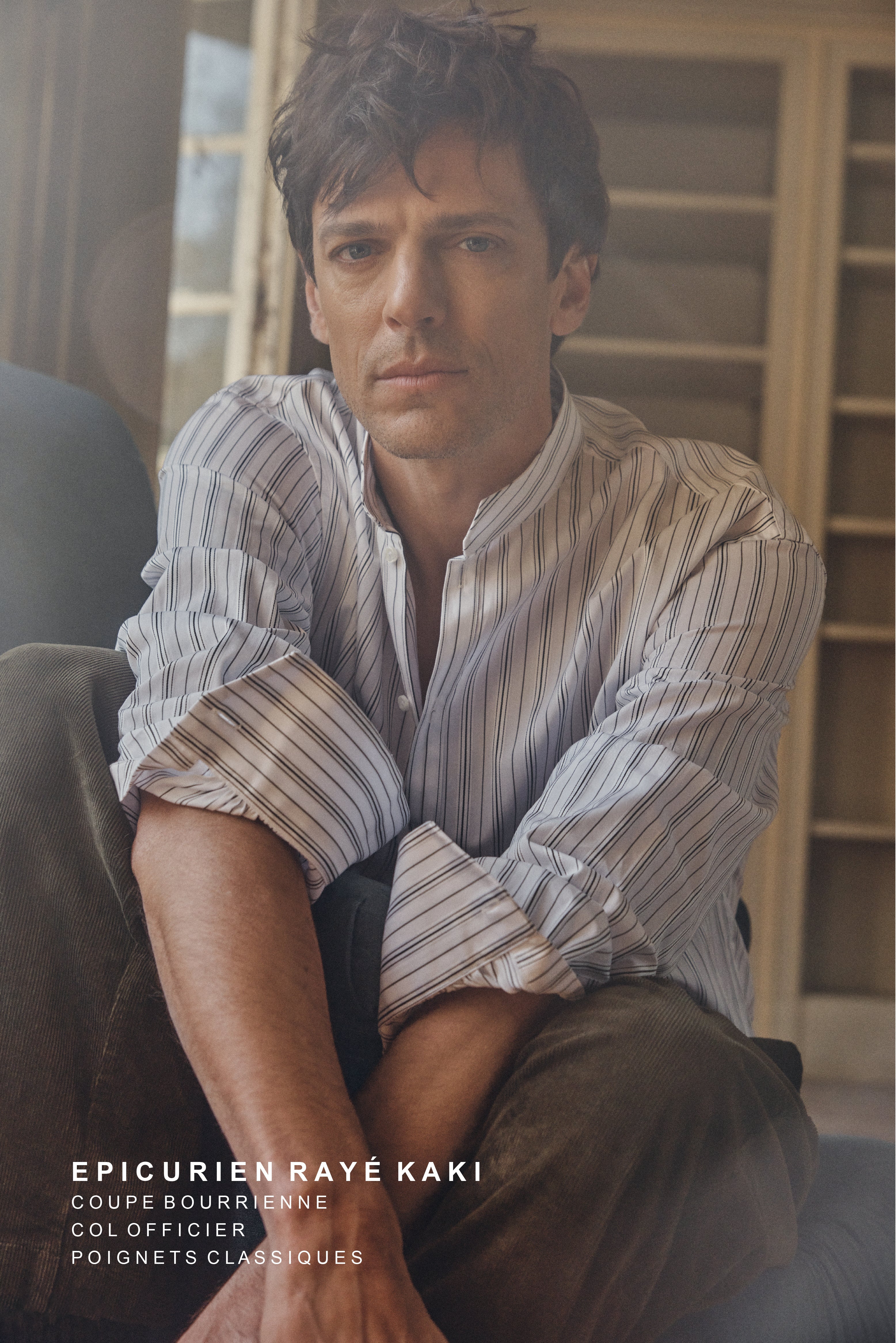 Modèle masculin assis de trois-quarts dans un intérieur chaleureux, portant la chemise ÉPICURIEN Rayée Kaki en popeline de coton : coupe ample Bourrienne, col officier biseauté et poignets classiques. Le tissu à fines rayures kaki capte la lumière douce, tandis que ses manches retroussées et son regard pensif évoquent une élégance décontractée.
