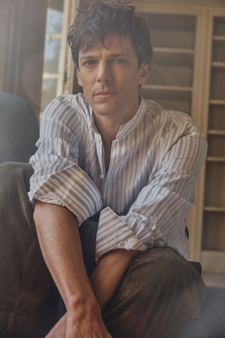 Modèle masculin assis dans un fauteuil, portant la chemise ÉPICURIEN Rayée Kaki en popeline de coton : coupe ample Bourrienne, col officier biseauté et poignets classiques. Le tissu à fines rayures kaki capte la lumière douce, tandis que ses manches retroussées et son regard pensif évoquent une élégance décontractée.