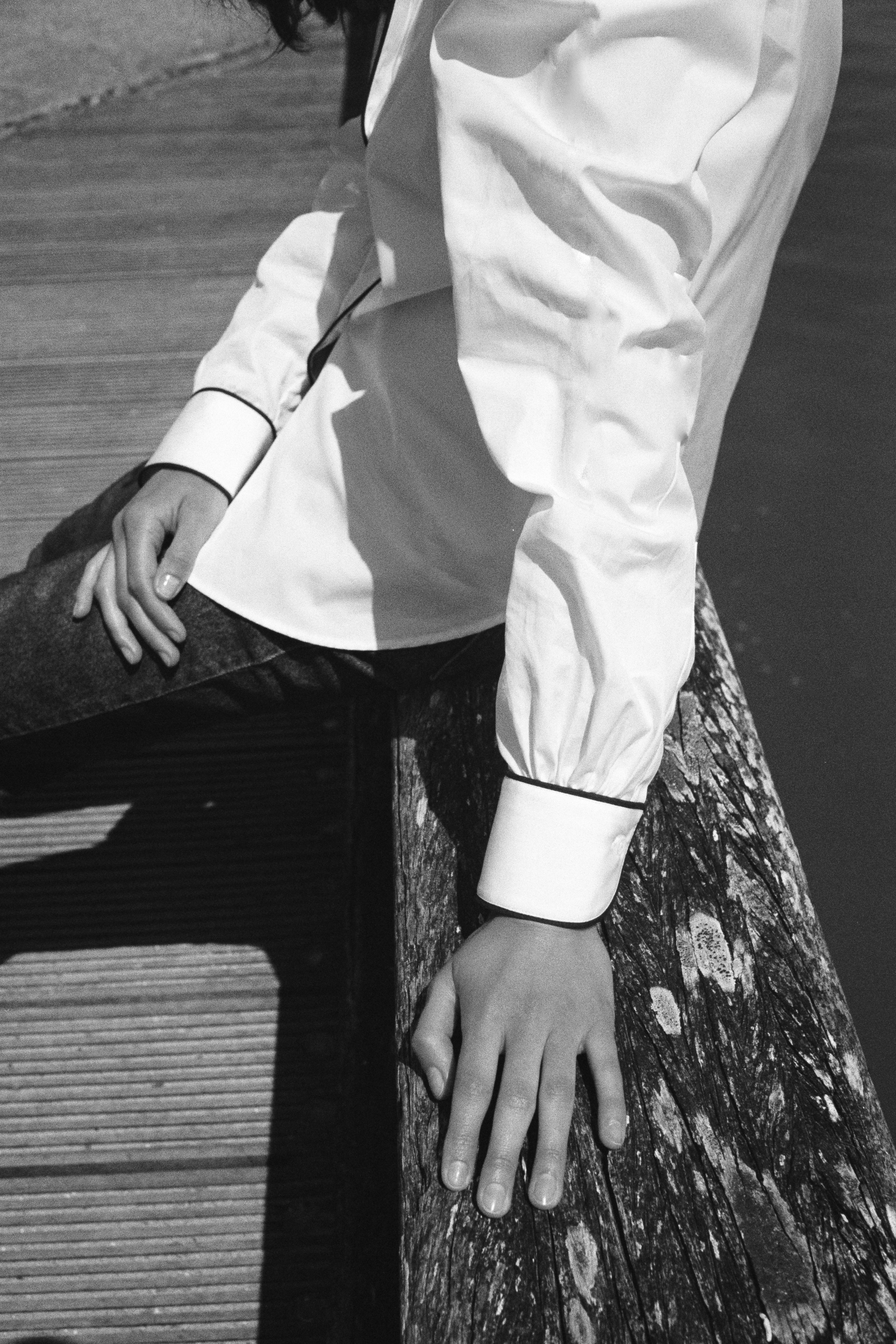 BOURRIENNE PARIS X: mannequin assise sur un muret, photo en noir et blanc, vue poignet, la chemise est conçue avec une popeline blanche et avec un passepoil bleu marine le long de la gorge de boutonnage ainsi que sur les poignets, les poignets ont des légères fronces.