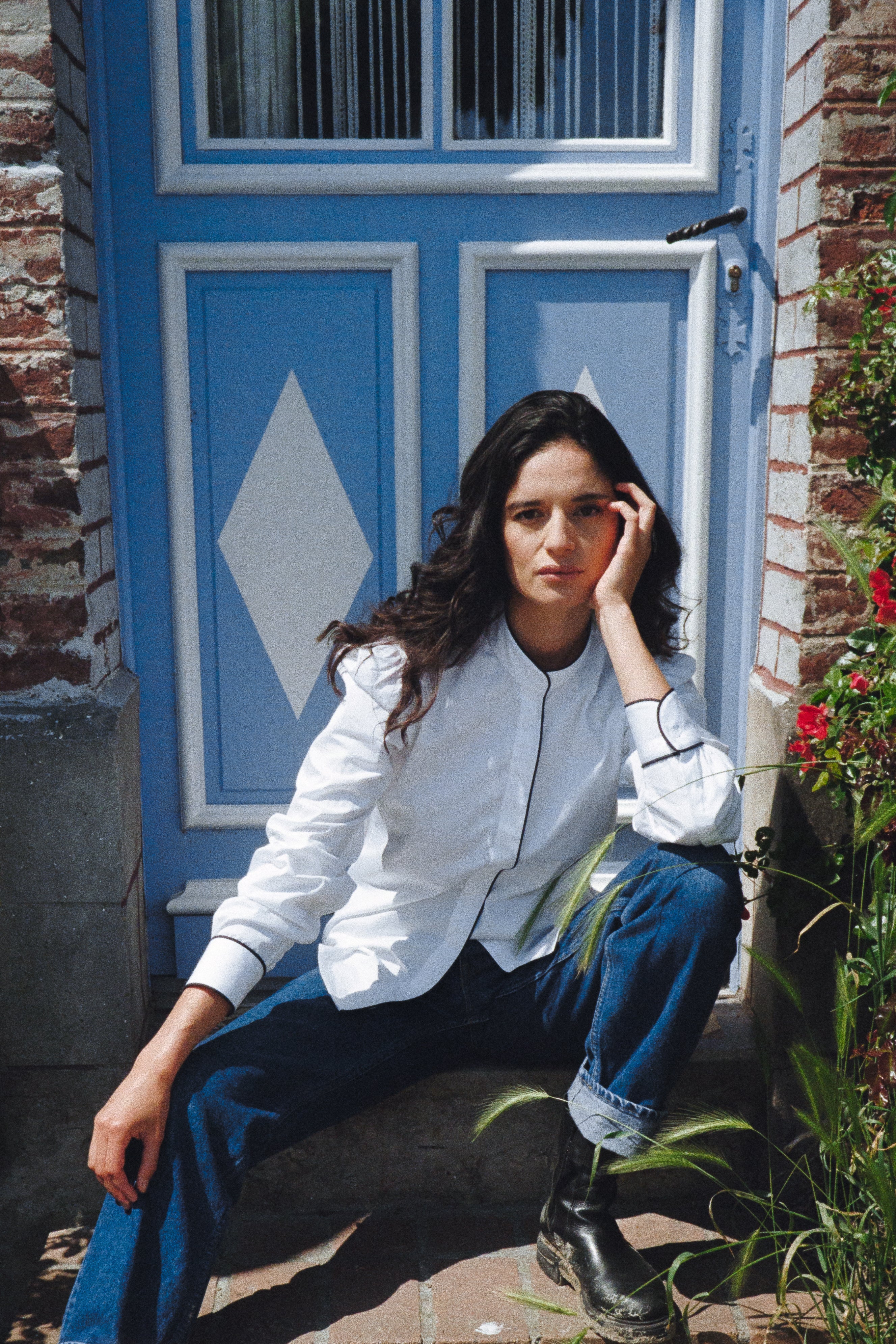 BOURRIENNE PARIS X: Mannequin assise sur une marche devant une porte, chemise est faite d'une popeline blanche ainsi que d'un passepoil bleu marine le long de la gorge de boutonnage ainsi que sur les poignets.