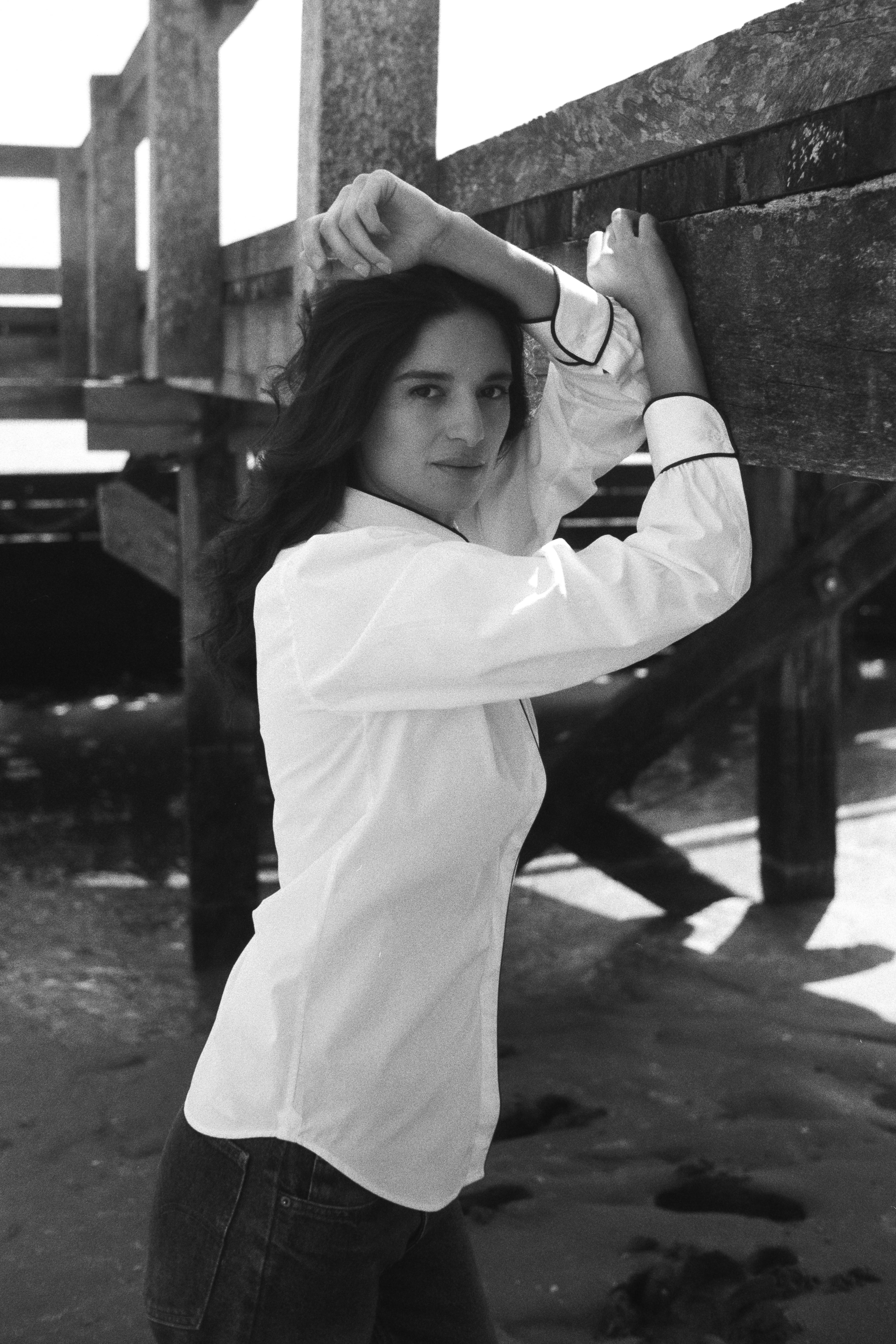 BOURRIENNE PARIS X: mannequin s'appuie contre un mur afin de mettre en avant les détails des poignets de la chemise blanche, liseré bleu marine sur les poignets, la photo est en noir et blanc.