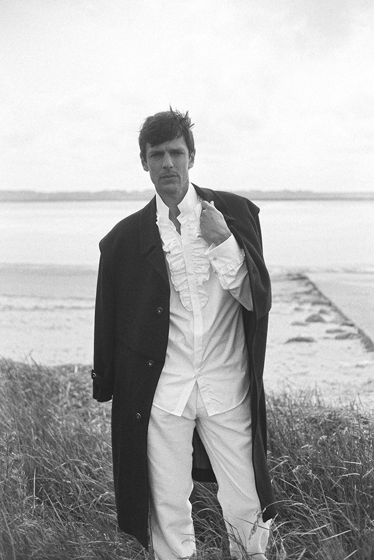 Photo prise de face, le mannequin porte une chemise en popeline blanche avec de nombreux détails à volants sur le devant de la chemise ainsi que sur les poignets. Il porte aussi un long manteau bleu marine sur les épaules.