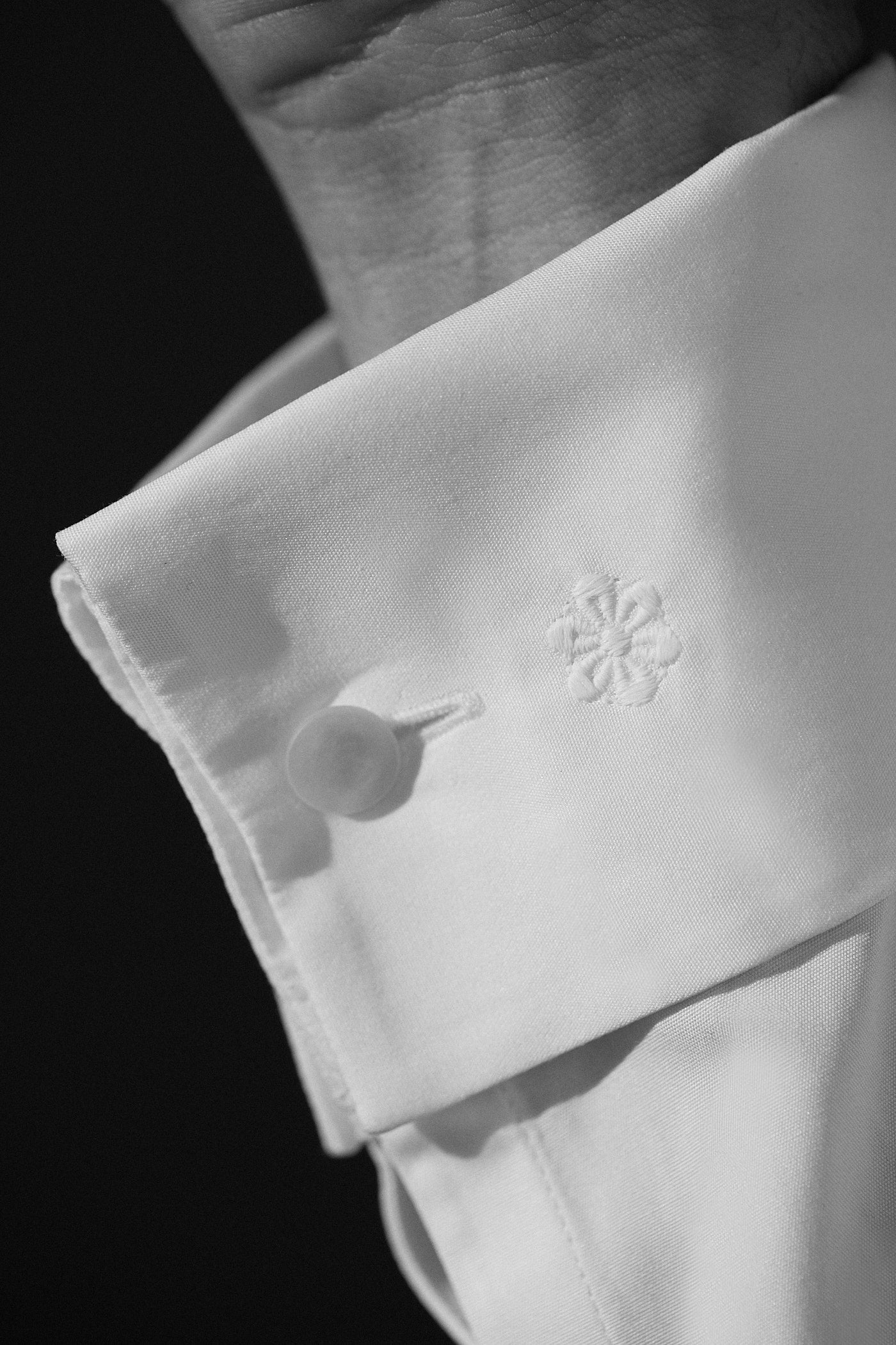 Photo noir et blanc plan serré d'un poignet d'homme, cet avant-bras est aperçu d'une chemise blanche en popeline blanche avec un poignet mousquetaire, on y voit la fleur Bourrienne brodée en blanc.