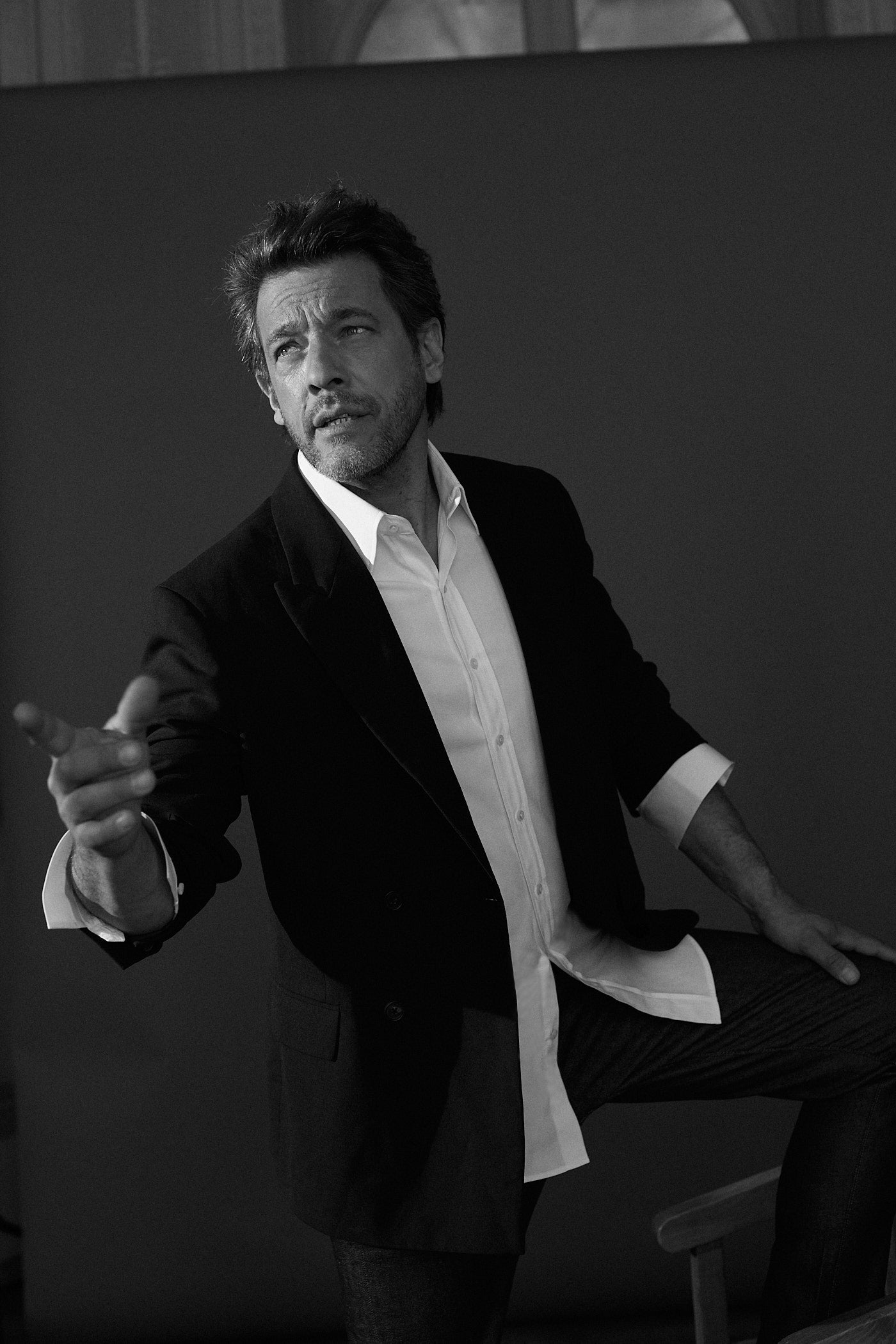 Photo noir et blanc plan américian d'un homme portant une chemise en popeline blanche double retors, col français et poignets bourrienne, une veste de costard noire et un jean. L'homme est debout, son pied gauche est élevé car il posé son pied sur une chaise. Sa main gauche touche sa jambe gauche et son bras droit pointe avec sa main quelque chose à sa droite. Son regard est tourné à sa droite.