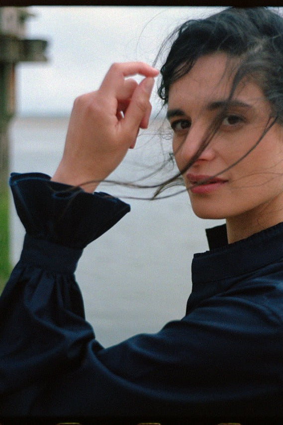 BOURRIENNE PARIS X: Photo de profil, le mannequin à des mèches de cheveux sur son visage et montre son avant bras de manière à ce qu’on puisse apercevoir les détails des manches bouffantes et des volants brodés au niveau des poignets.