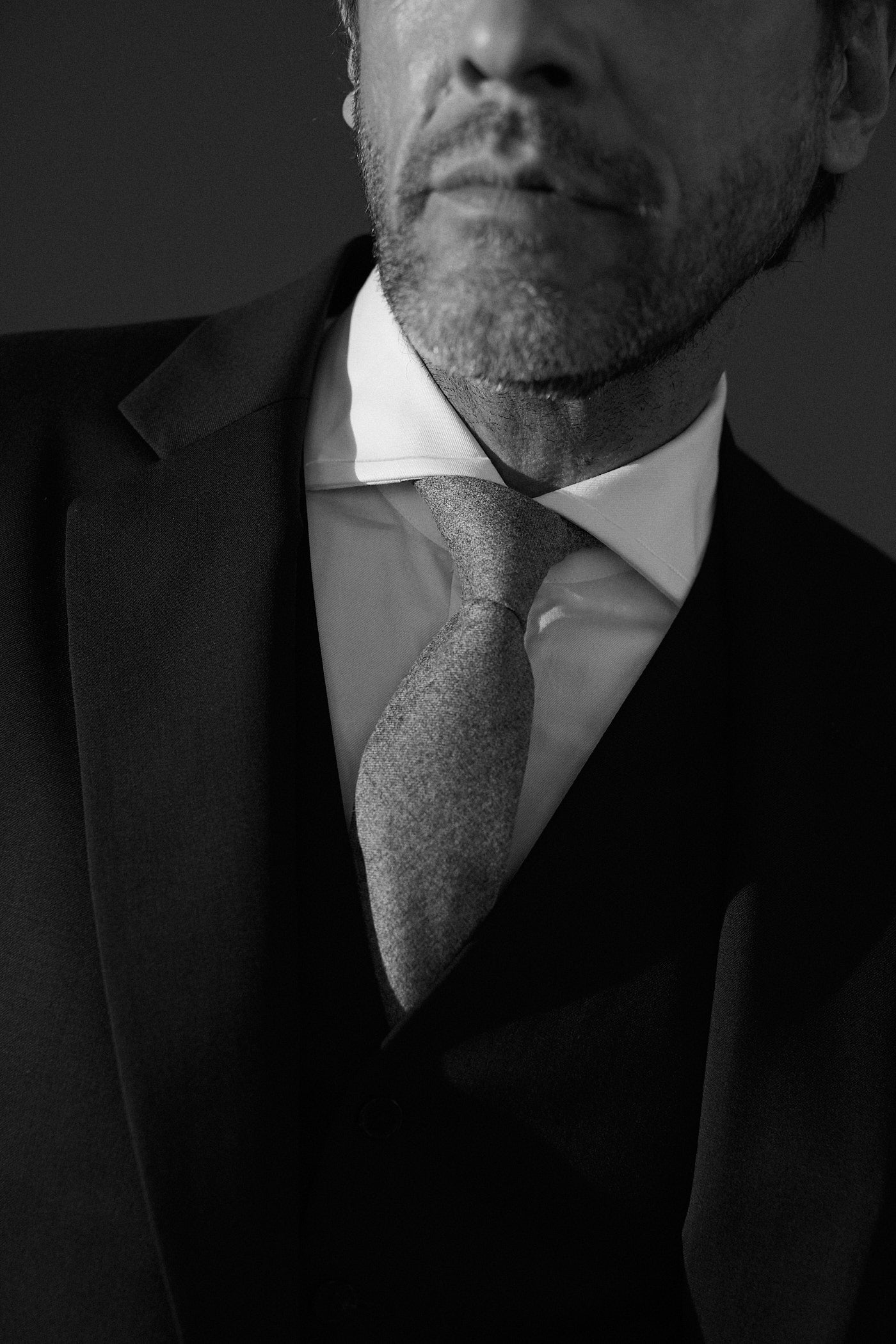 Photo noir et blanc plan américain, d'un homme portant un costard 3 pièces, veste noire et chemise blanche en popeline double retors avec un col italien, une cravate grise nouée au col de la chemise. On aperçoit au troisième plan son visage jusqu'au nez.