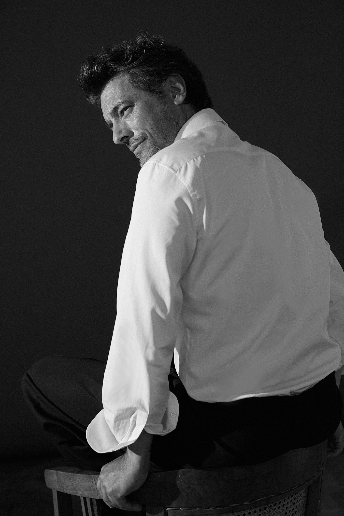 Photo en noir et blanc plan américain. Un homme est tourné de dos il est assis sur le dossier d'une chaise. Il porte une chemise blanche en popeline double retors, col italien et poignets classiques. on voit en