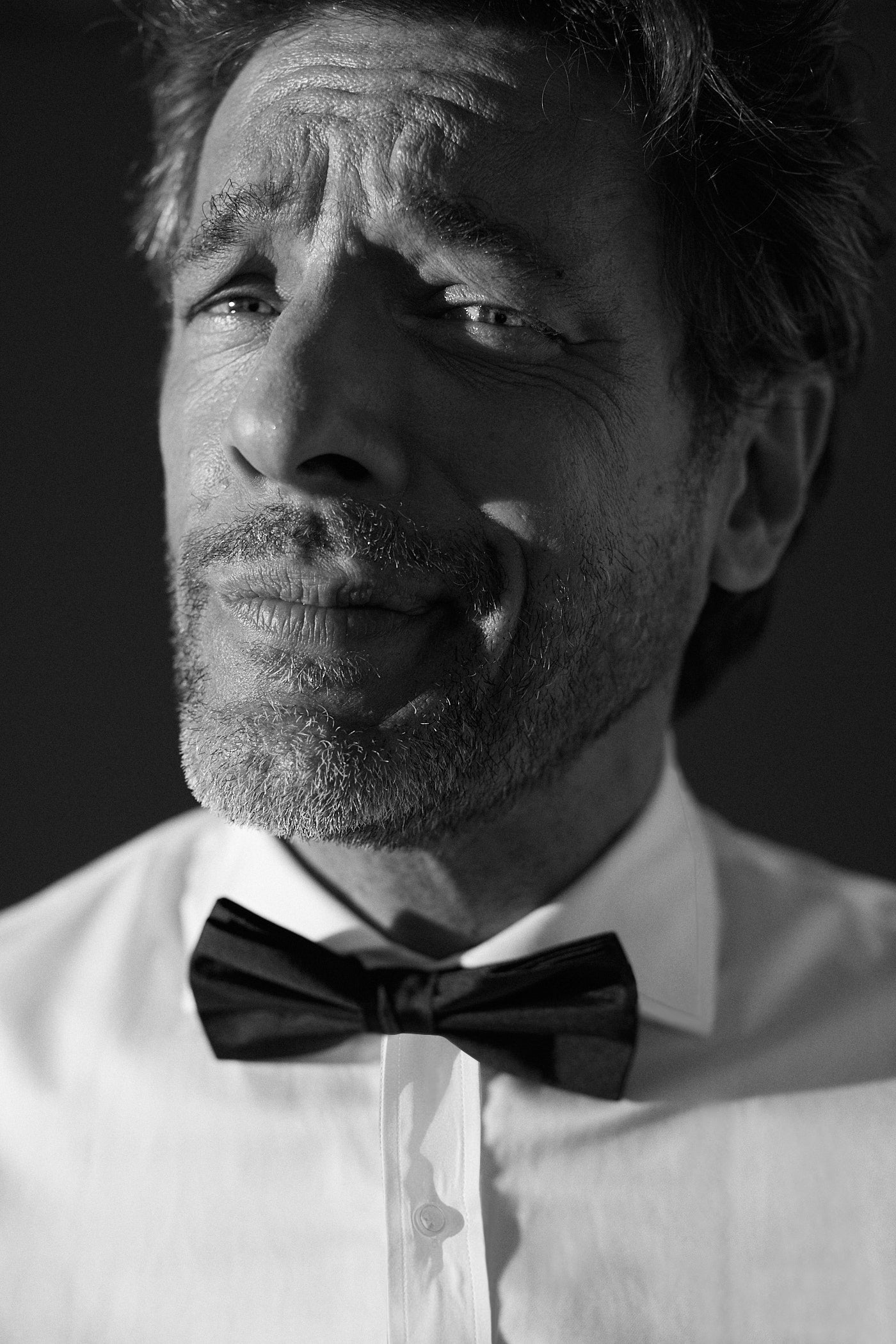 Photo en noir et blanc gros plan d'un homme portant une chemise blanche en popeline double retors avec un col Italien. Le col est noué avec un noeud papillon noir au premier plan, au deuxième plan le visage de l'homme qui regarde l'objectif, il sourit sans montrer ses dents, la bouche part vers la gauche et crée un plis sur sa joue gauche.