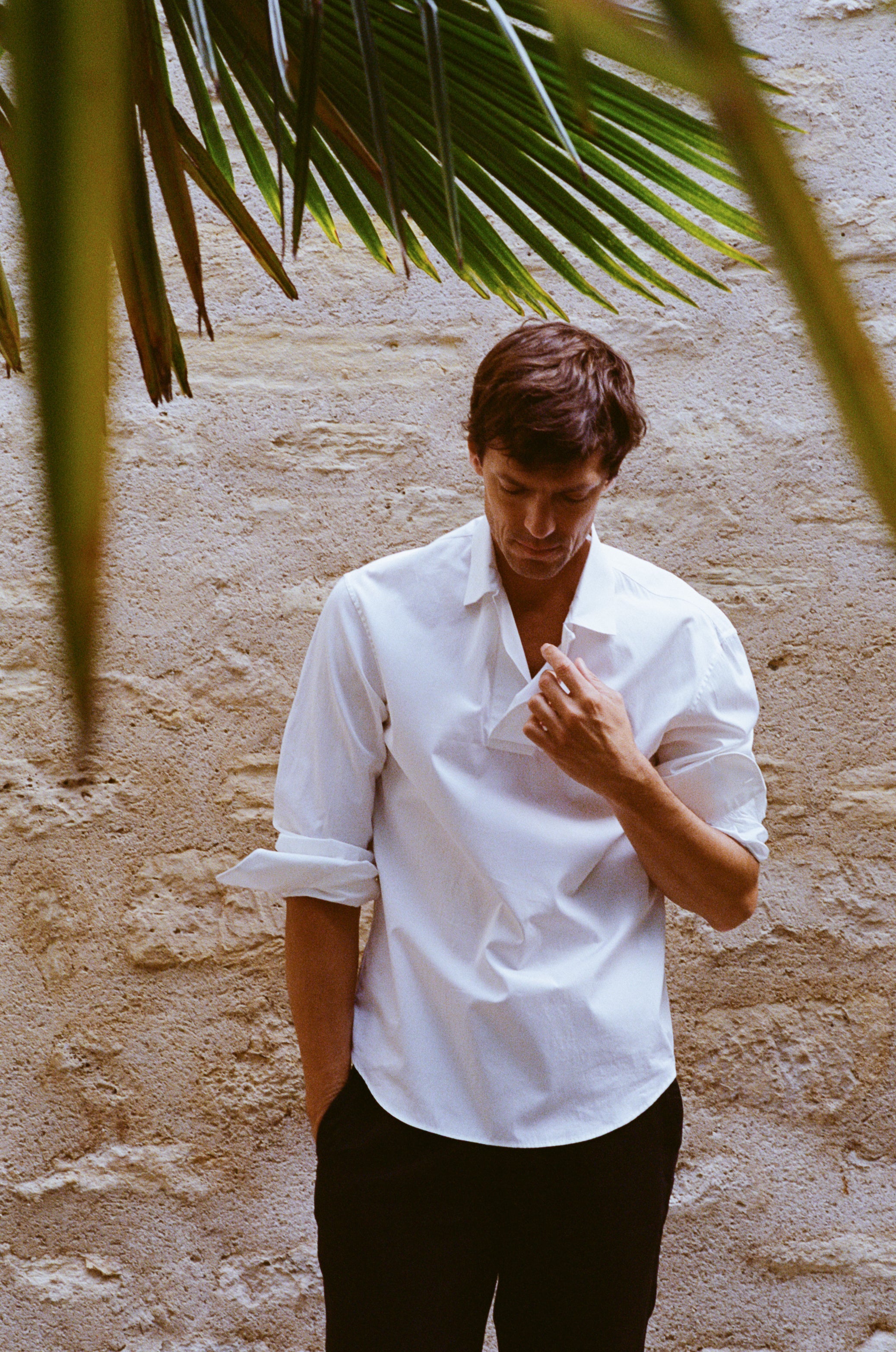 Homme portant une chemise blanche élégante aux manches retroussées, photographié en extérieur devant un mur en pierre.