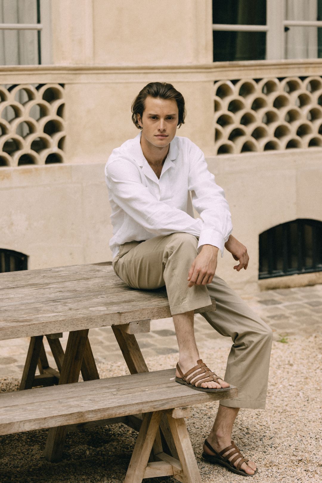 Homme assis sur une table en bois en extérieur, vêtu de la chemise blanche en lin Marin de Bourrienne Paris X et d’un pantalon beige, allure décontractée.