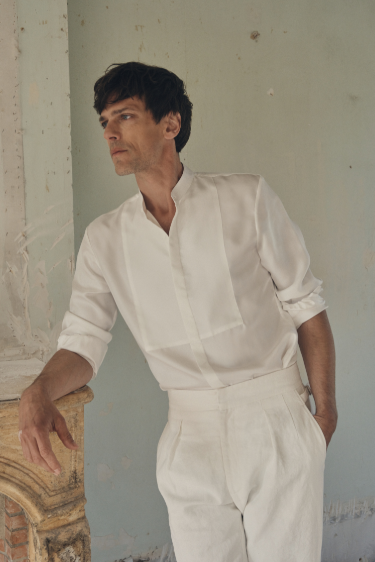 Homme de profil trois-quarts, appuyé contre une cheminée en pierre patinée, portant une chemise NOCTURNE en soie blanche à coupe ajustée, col officier et plastron revisité. Les manches sont retroussées au-dessus du poignet et le plastron en nid d’abeille ajoute un relief subtil sur la poitrine. Le décor minimaliste aux murs clairsemés et la lumière naturelle mettent en valeur la fluidité et la finesse du tissu.