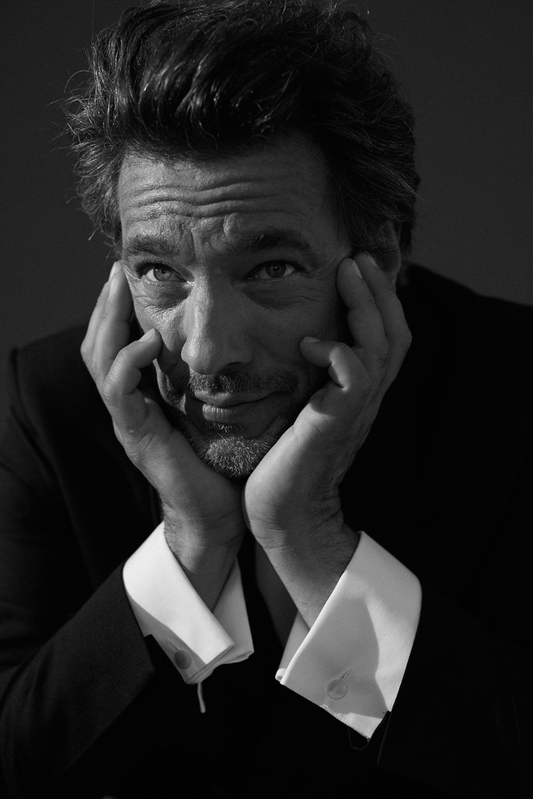 Photo noir et blanc plan serré d'un homme qui tient sa tête avec ses deux mains. Il porte une chemise blanche en popeline blanche double retors, poignets mousquetaires, avec une veste costard noire. Il regarde à sa gauche, l'air pensif.