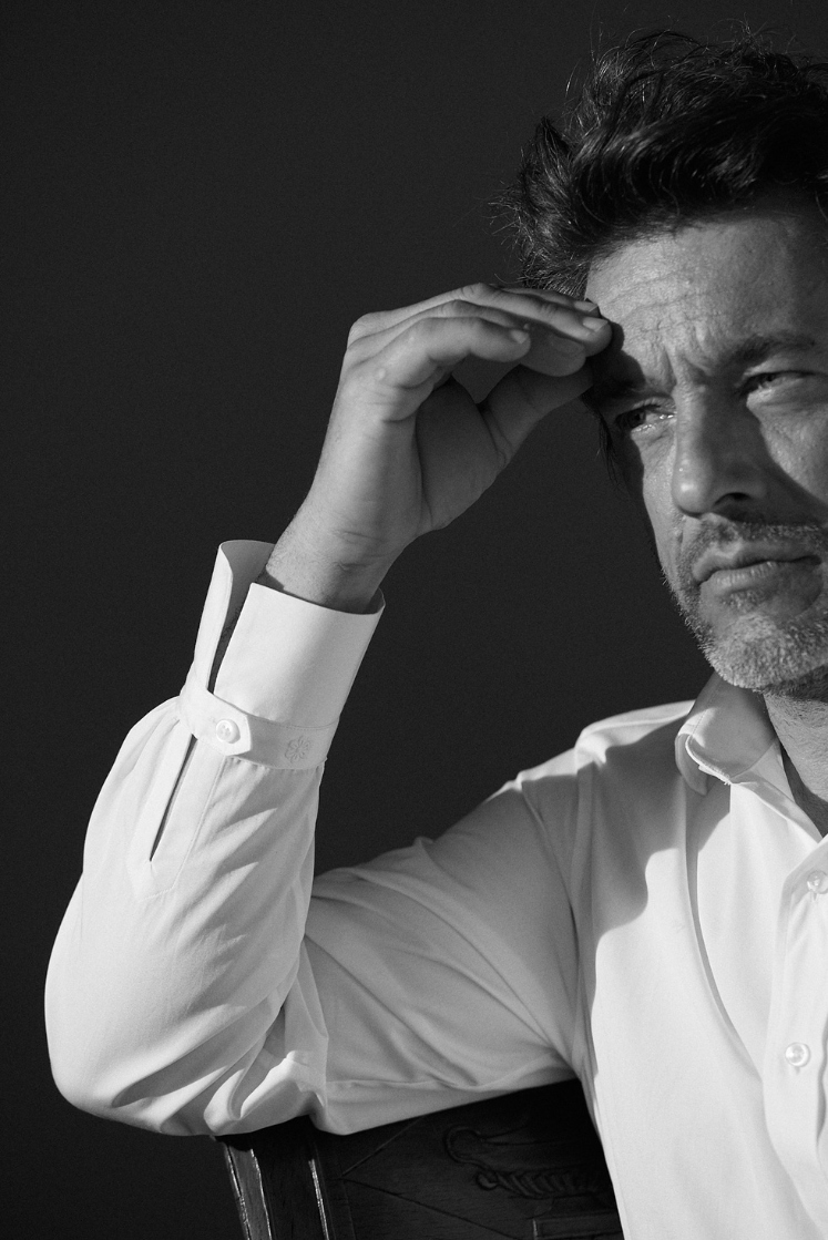 Photo plan poitrine en noir et blanc d'un homme assis portant une chemise blanche col italien et poignet Bourrienne. On aperçoit seulement son bras droit qui est posé sur le haut de dossier de sa chaise. Le bras est plié, sa main touche son front, on voit le poignet Bourrienne. Le regard de l'homme est profond il regarde à sa droite.