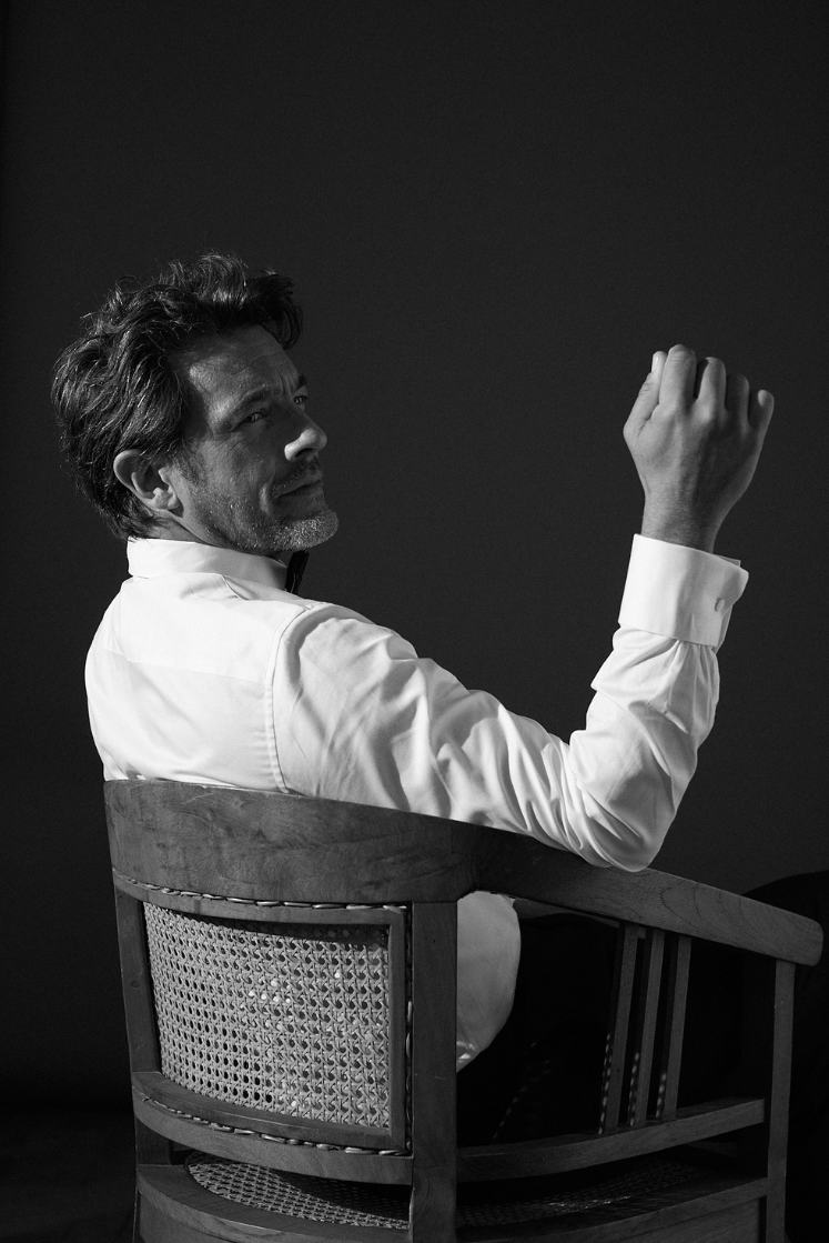 homme portant une chemise blanche avec un col italien, il est assis de profil sur une chaise en osiers, son visage est tourné vers la droite, il pose sont coude sur l’accoudoir de droite, la main vers le haut, on aperçoit le poignet mousquetaire de la chemise blanche