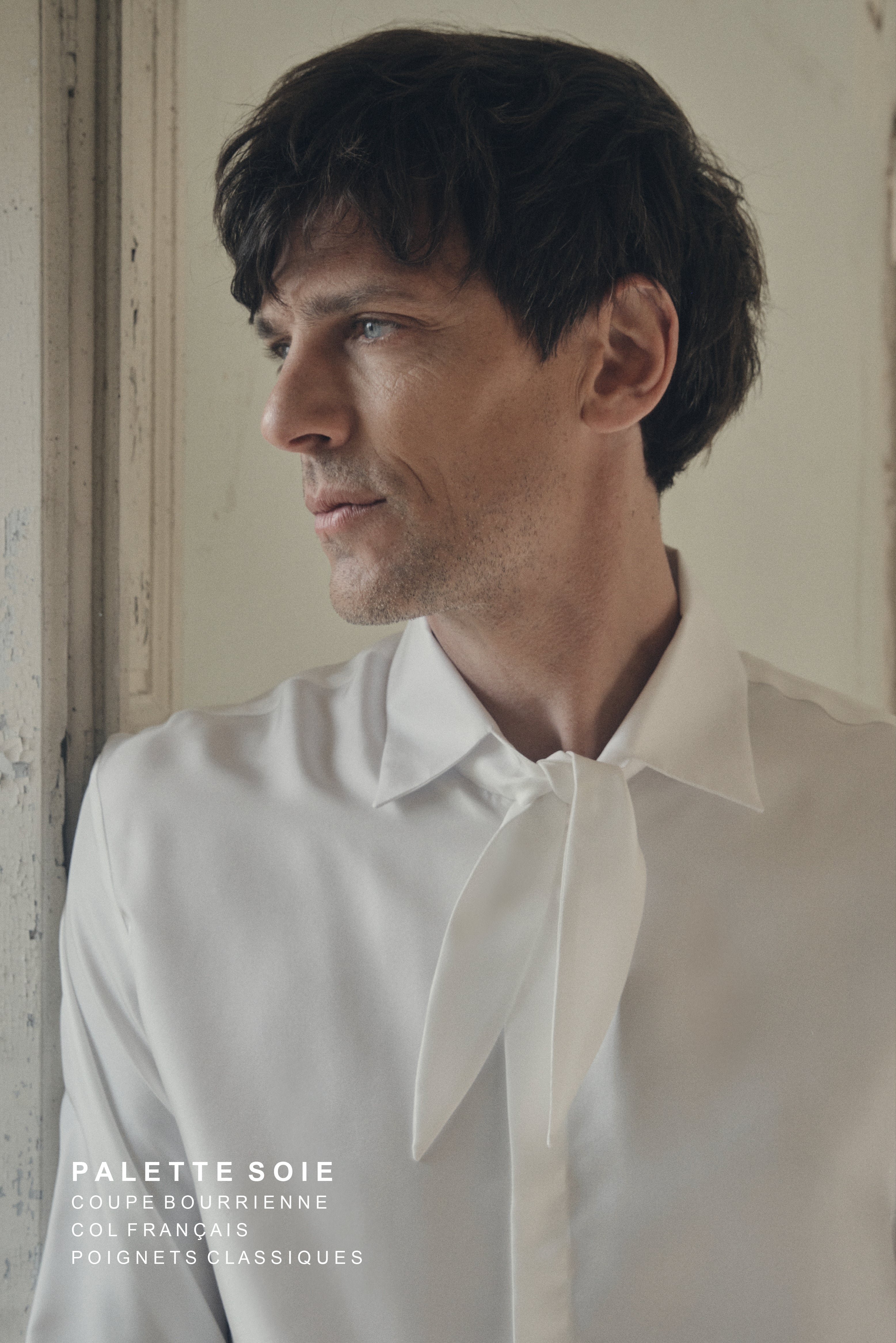 Homme de profil, appuyé contre l’embrasure d’une fenêtre, portant la chemise PALETTE en soie blanche à coupe Bourrienne et col français, dont le large nœud décontracté apporte une touche de fluidité. Le décolleté ouvert et le tombé souple du tissu mettent en valeur l’éclat satiné de la soie, tandis que les poignets classiques ajoutent une note raffinée. La lumière douce qui filtre par la fenêtre souligne la texture délicate du vêtement et le contraste entre l’élégance moderne et le décor patiné.