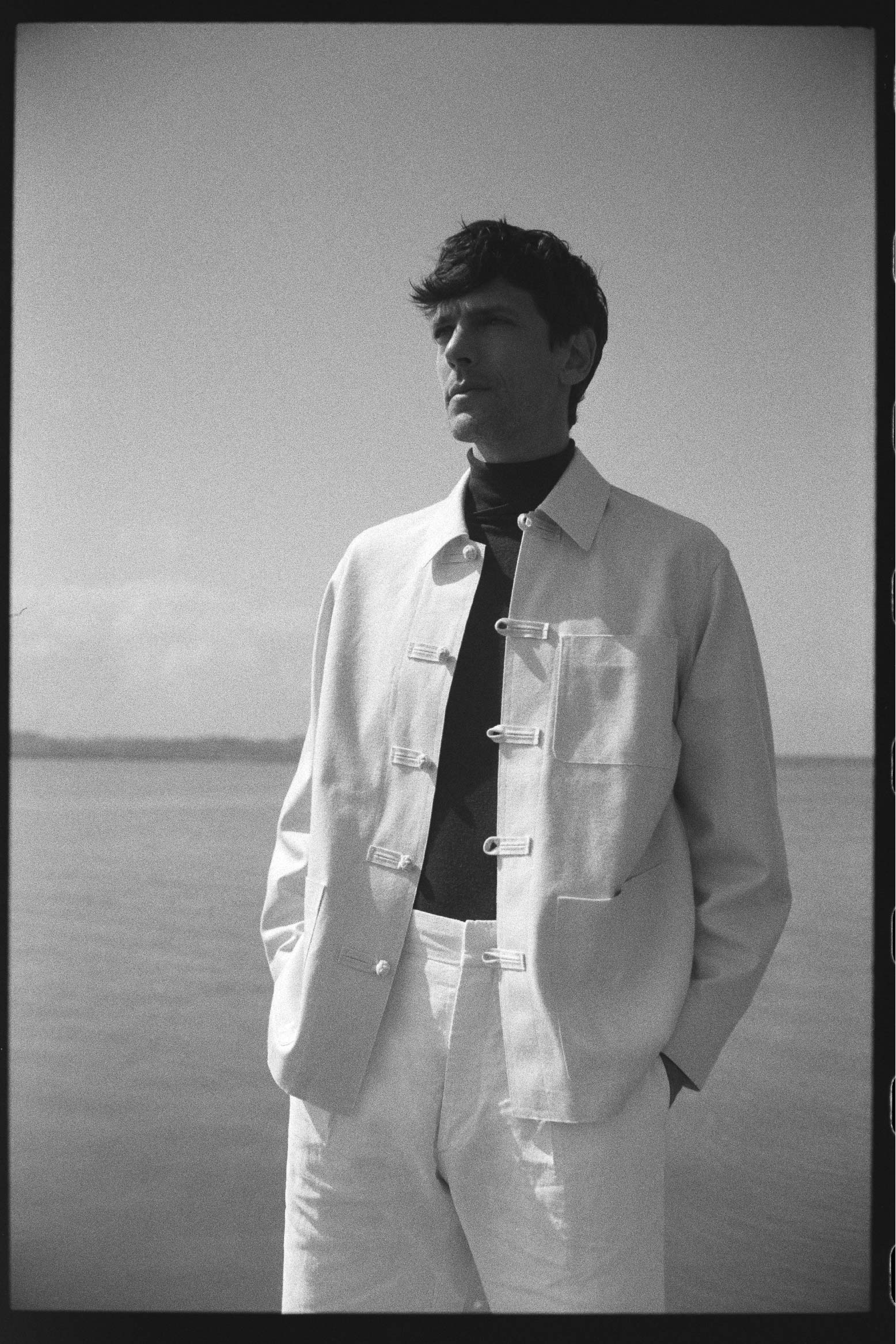 BOURRIENNE PARIS X: Photo prise de face, le mannequin est devant la plage et regarde à l’horizon, il a les mains dans les poches et porte une veste claire à col français et au boutonnage travaillé, disposant de trois poches plaquées à l'avant. La coupe est ajustée et les poignets sont classiques.