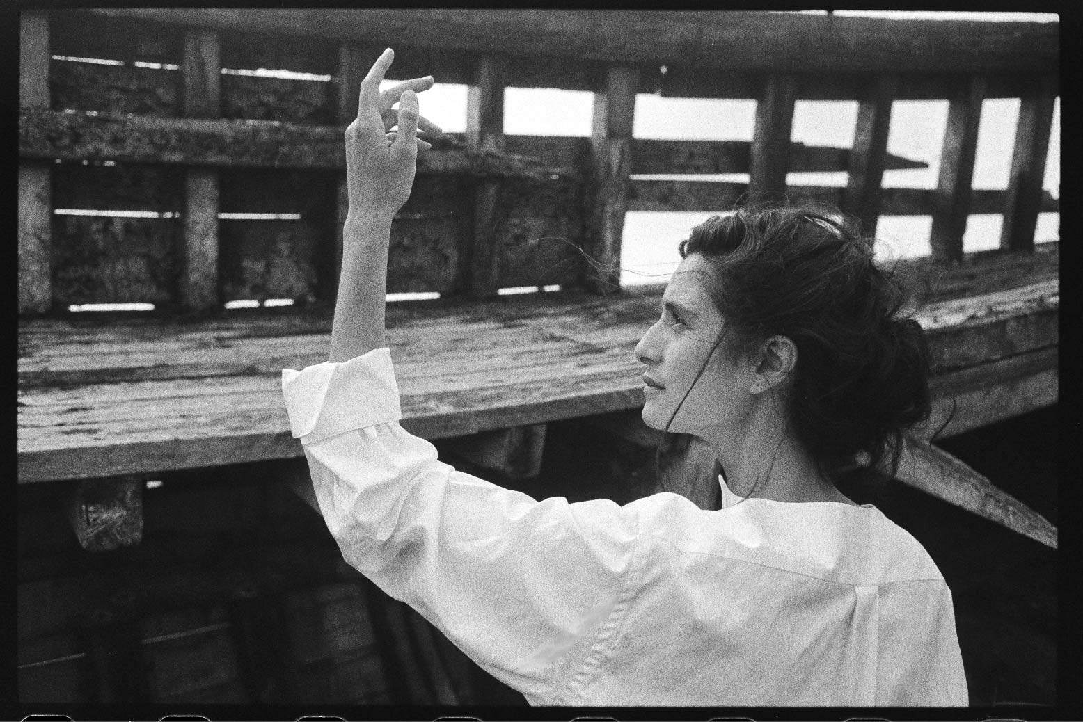 BOURRIENNE PARIS X: Photo prise de dos, le mannequin regarde sa main qui est levée vers la gauche. Elle porte une chemise blanche coupée dans une popeline de coton douce et de grande qualité d’origine japonaise. Le chemisier est non boutonné et a un décolleté fendu est orné de part et d'autre d'un plastron en ogive qui allonge la silhouette.